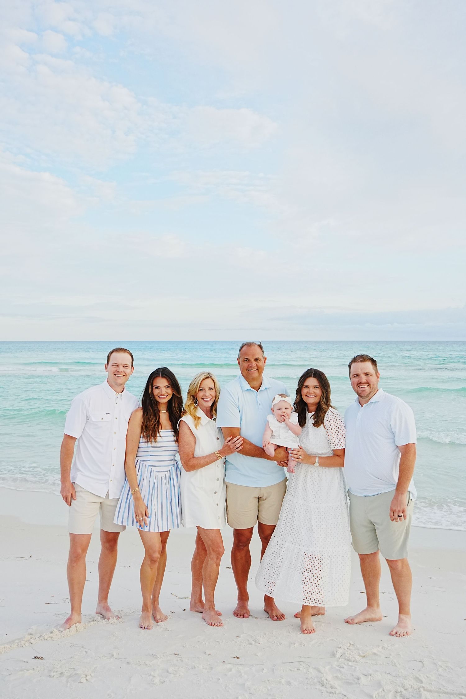 Multi-generational family wearing coordinated white and blue outfits poses together on sunny beach with turquoise water.