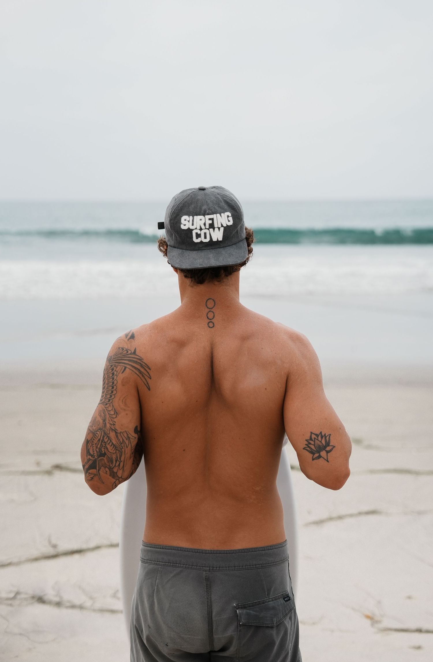 Person with tattoos wearing a baseball cap photographed from behind on a beach.
