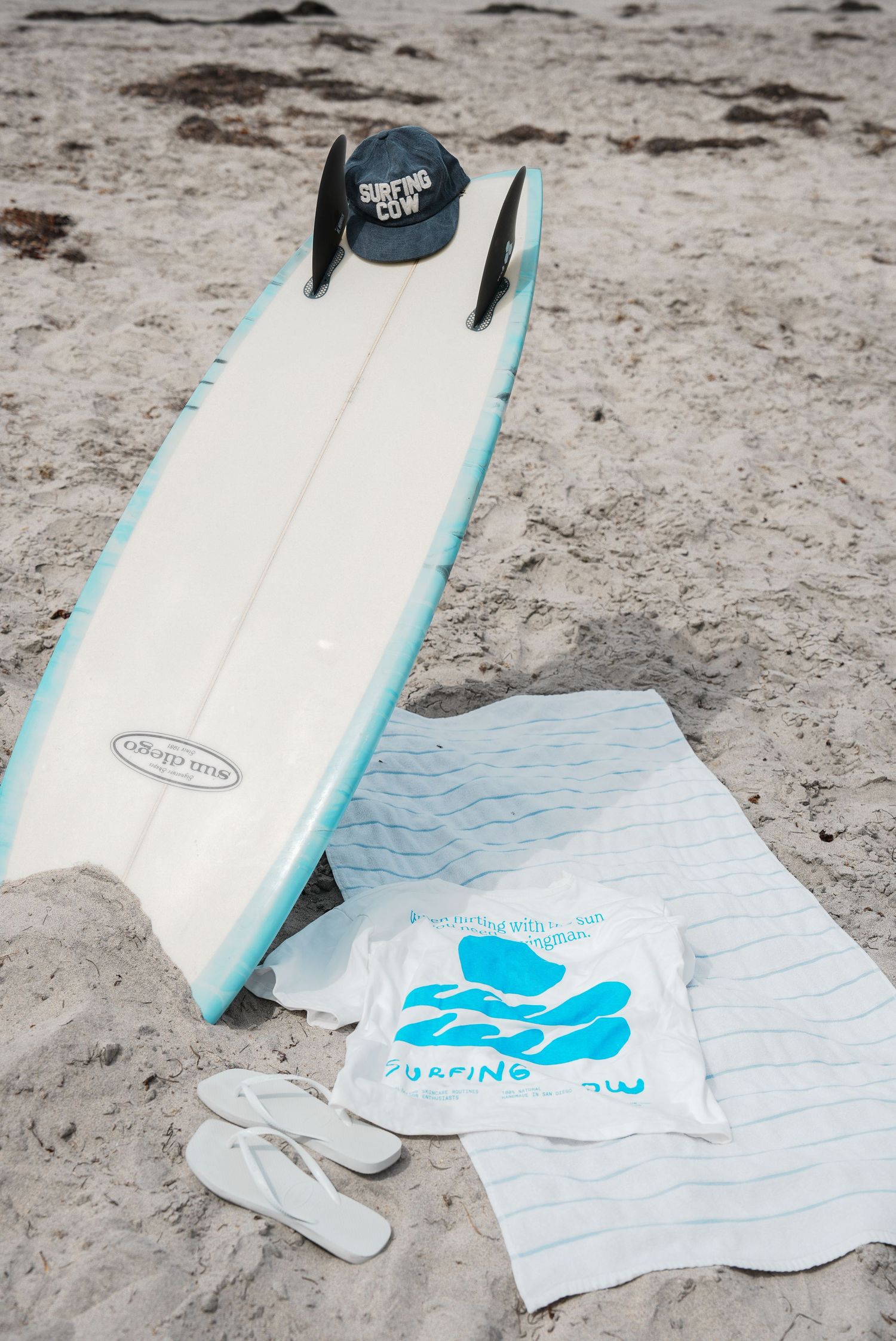 White surfboard with blue accents and logo displayed on beach towel against sandy backdrop.