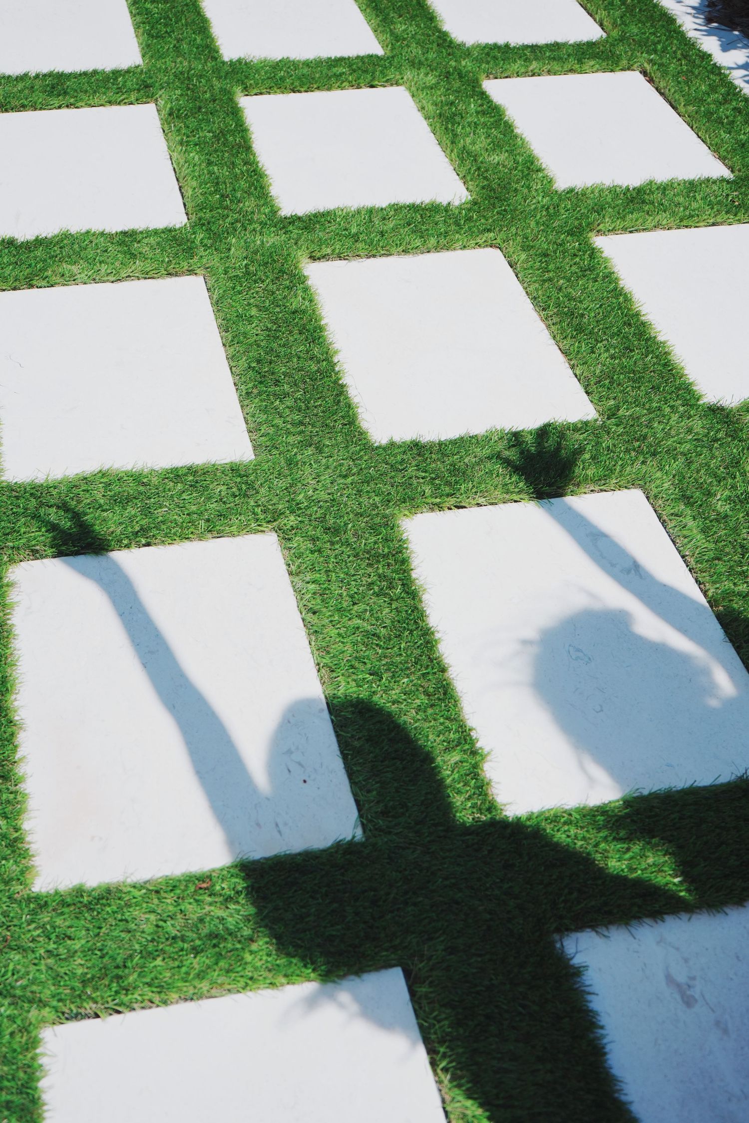 Shadows cast on geometric white pavers with green grass strips between them in outdoor garden design.