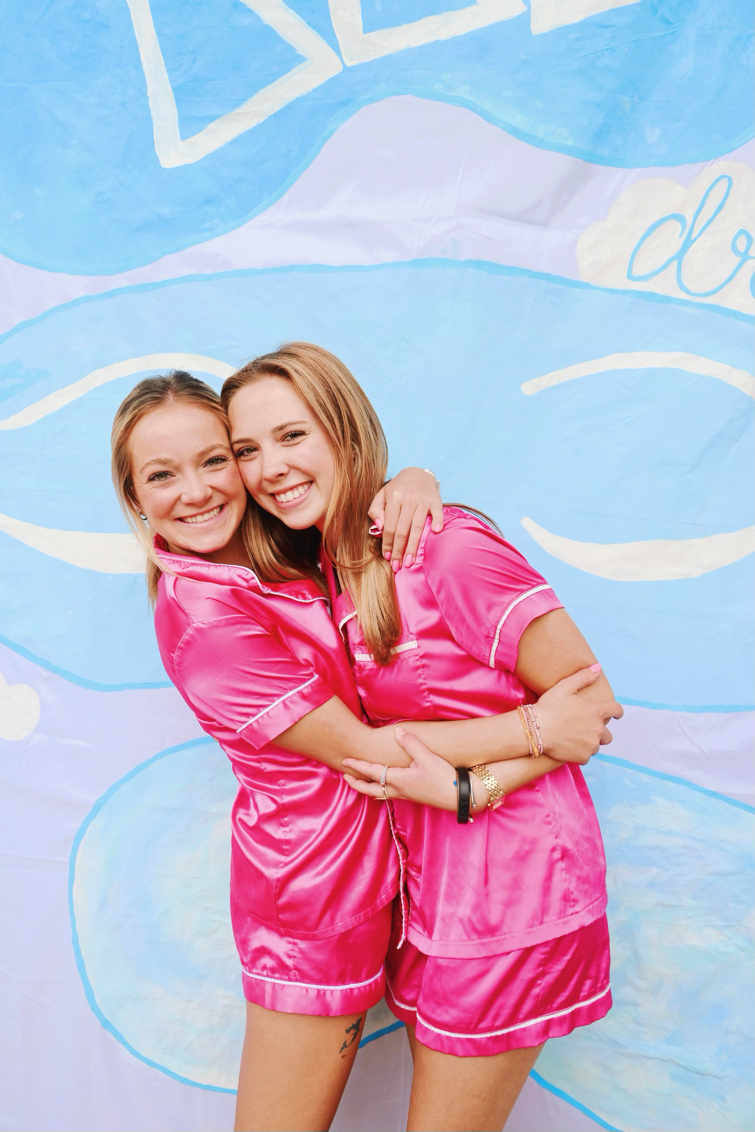Two people wearing matching pink satin pajama sets embrace and smile against a blue and white painted wall background.