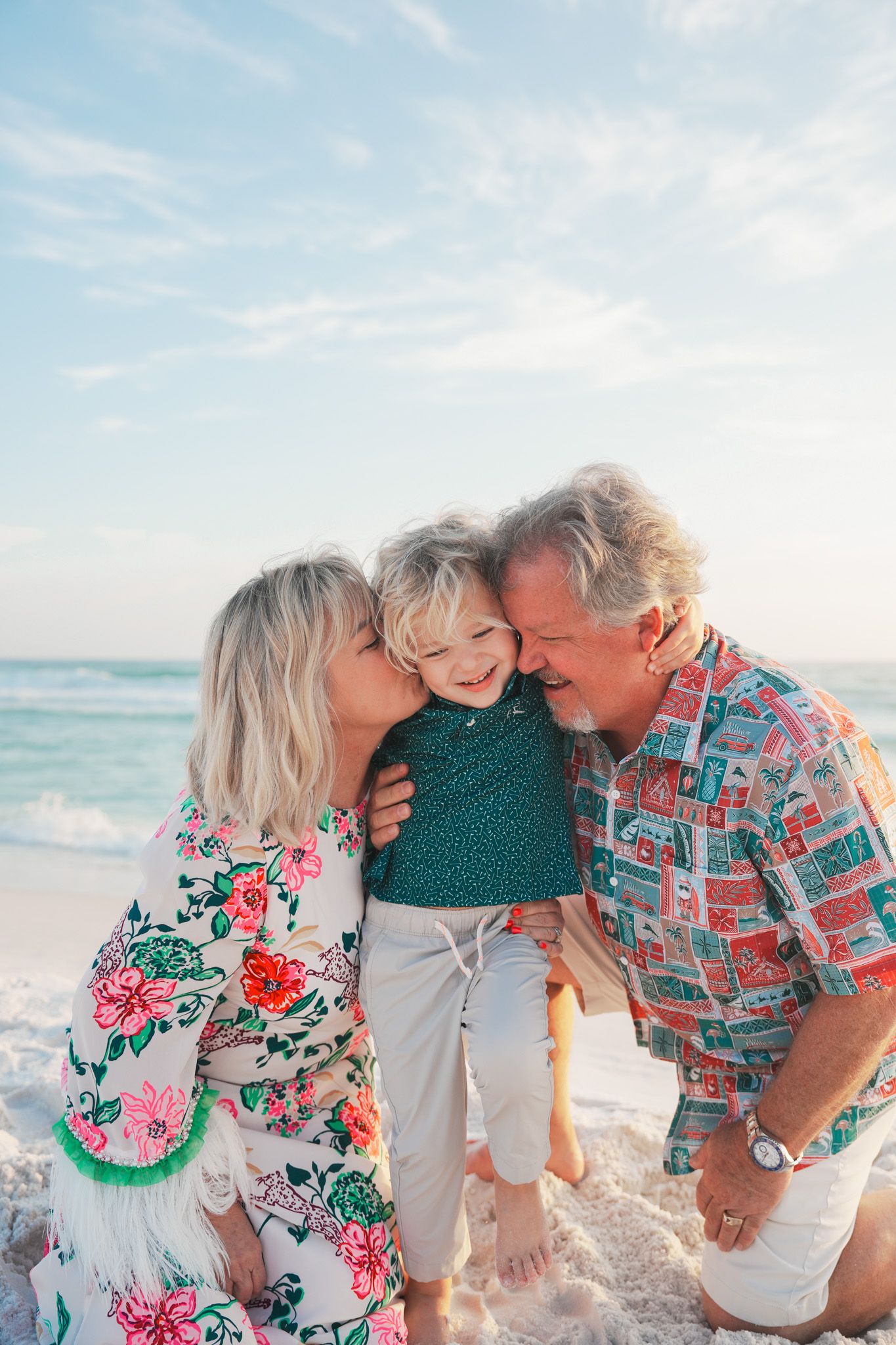 A joyful family moment at the beach with three people wearing floral and patterned outfits embracing in the sunshine.
