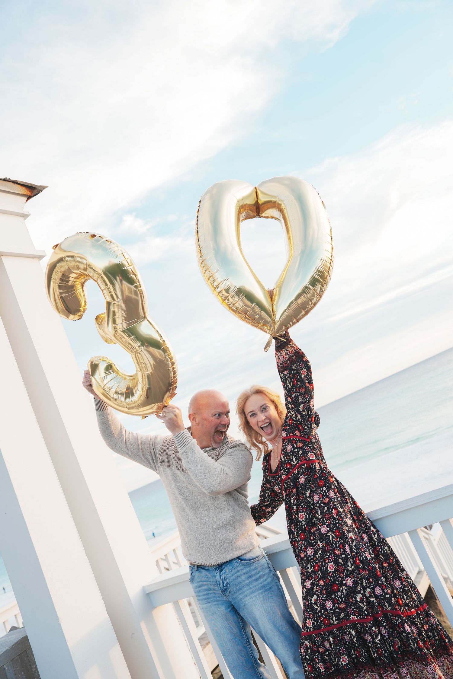 Two people hold silver balloon letters while celebrating together against a bright sky background.