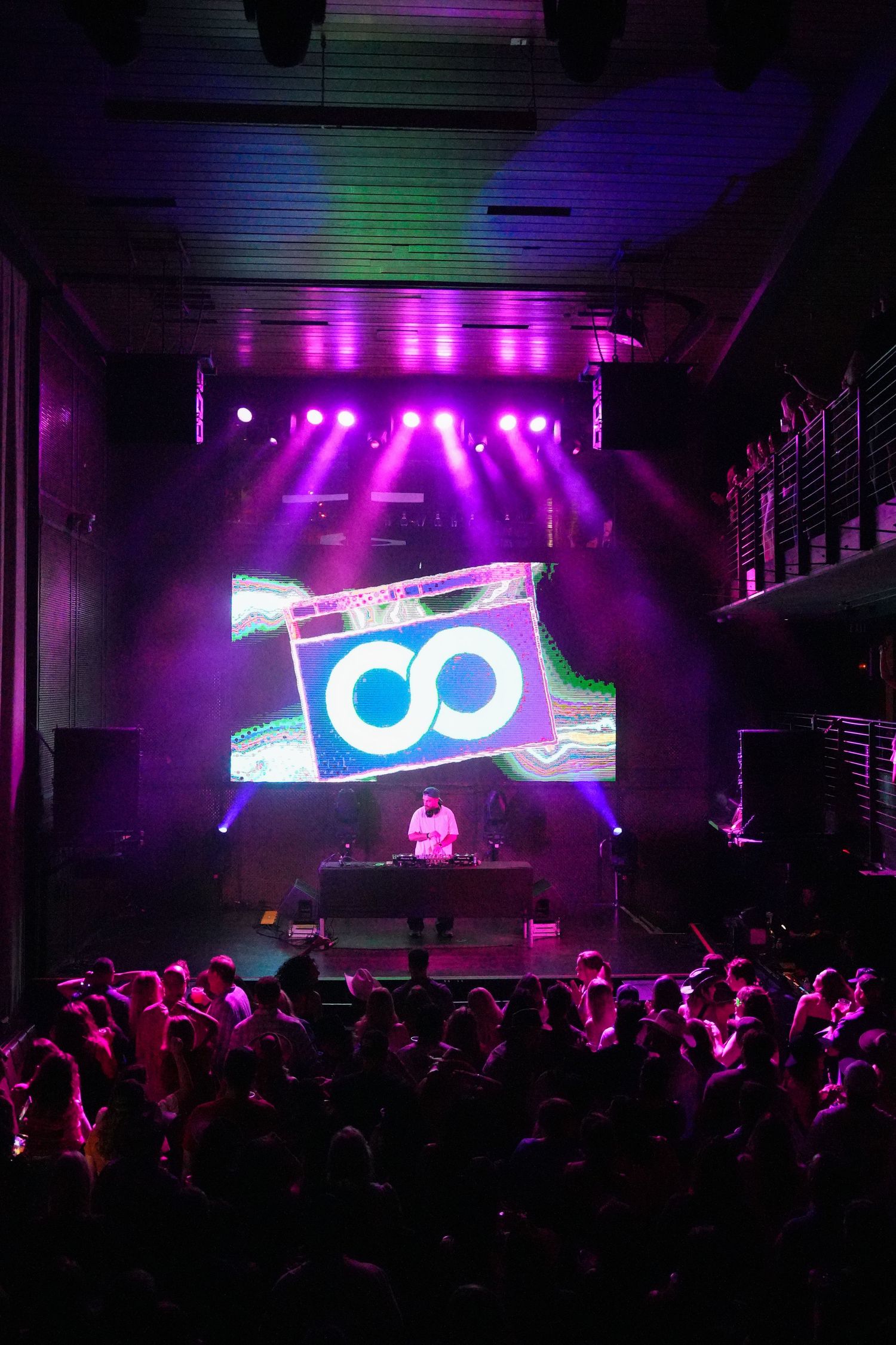 Concert crowd silhouettes against vibrant purple lighting and digital displays at an electronic music performance.