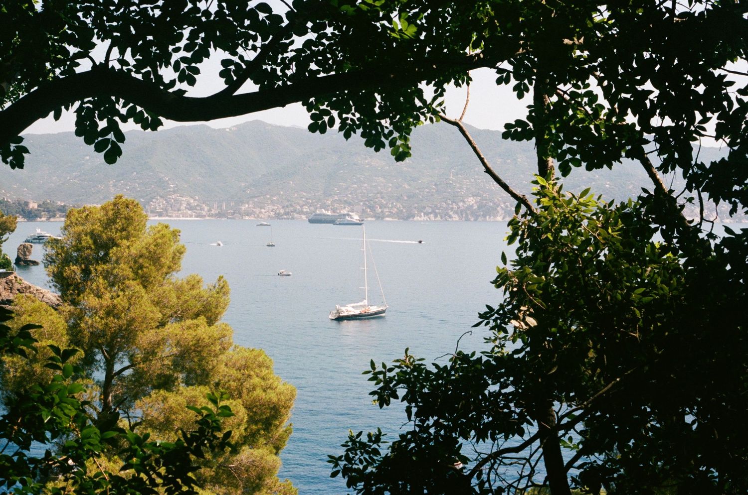 Sailboat anchored in calm blue Mediterranean bay framed by lush green trees with hillside village in background.