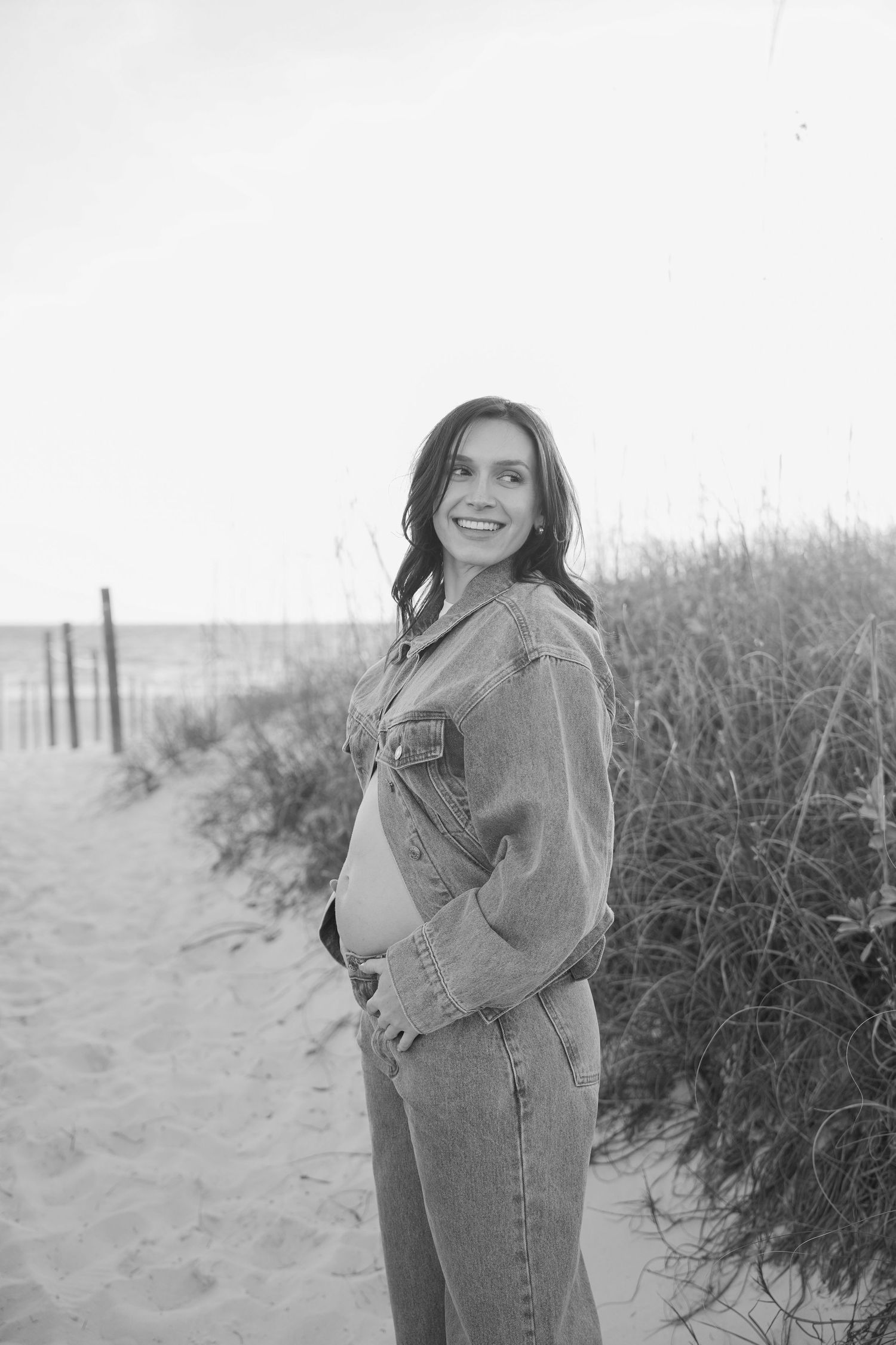Smiling pregnant woman in denim jacket and jeans poses on sandy beach path with sea grass in black and white photo.