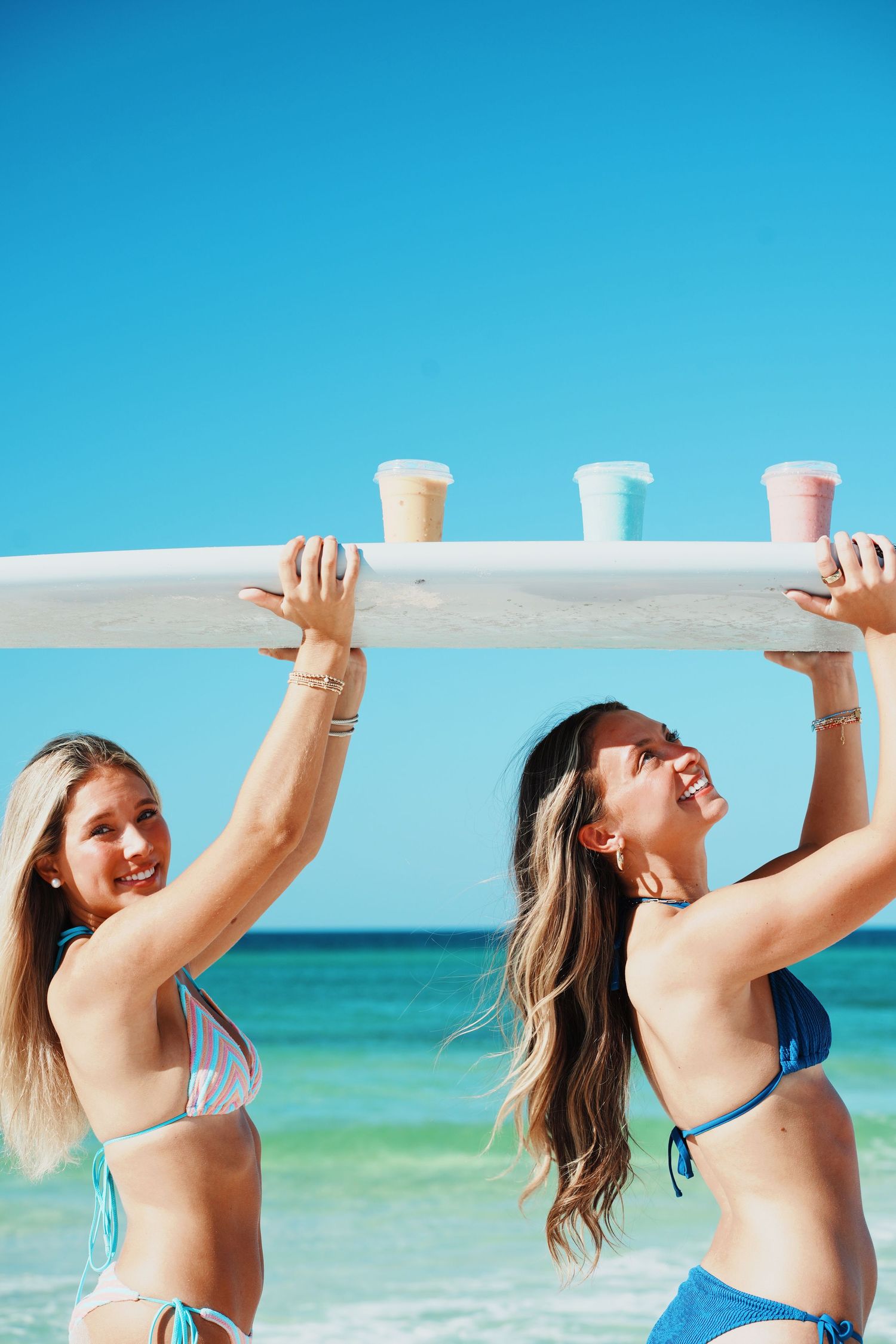 Two women in bikinis smiling and holding drinks on a beach bar while enjoying a sunny day with turquoise ocean backdrop.