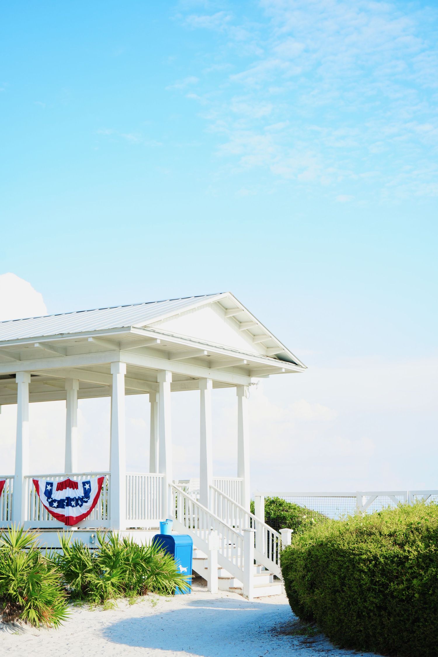 White coastal gazebo with patriotic bunting and palm plants along a curved pathway against bright blue summer sky.