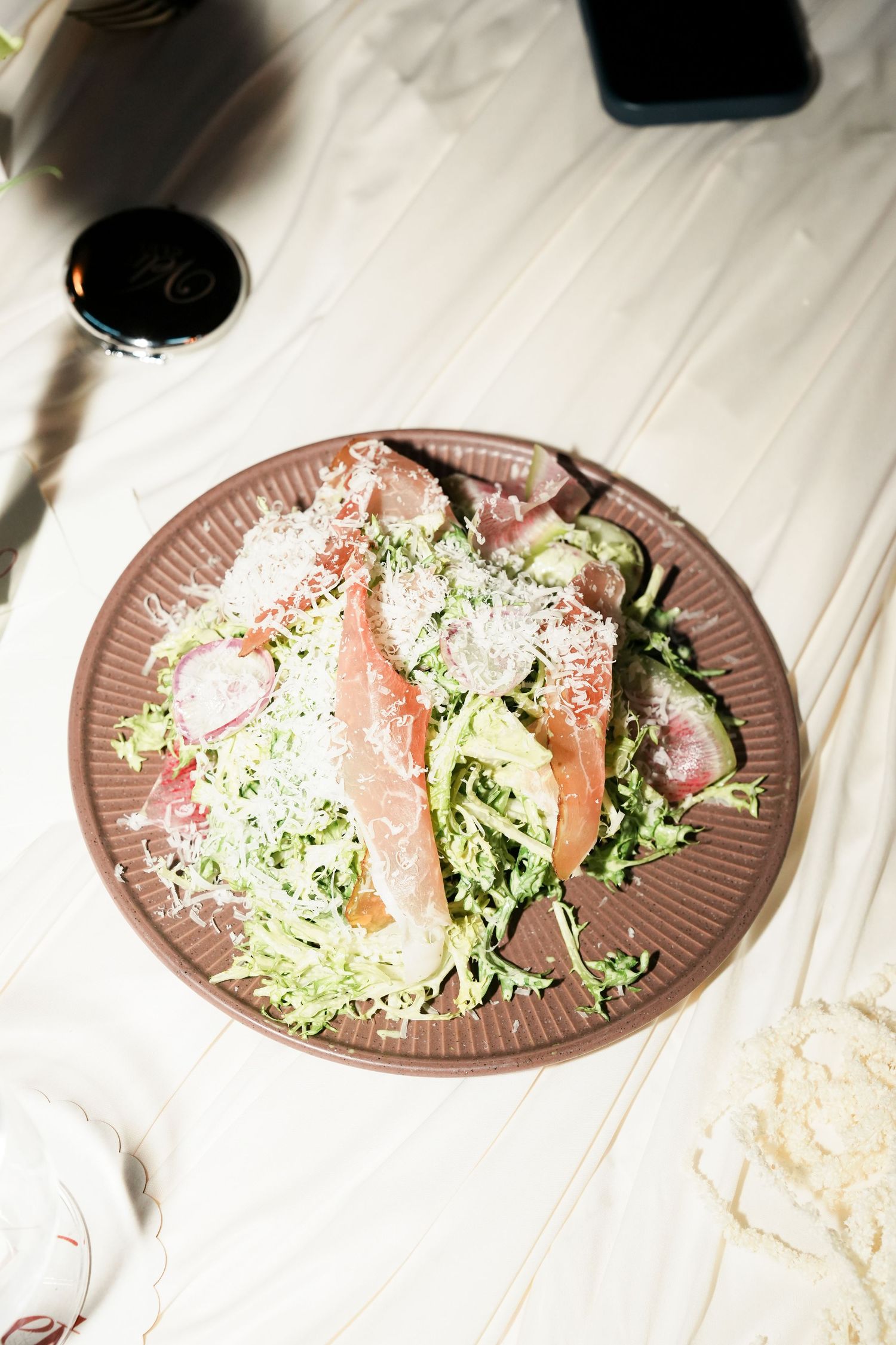 Fresh green salad with prosciutto, shaved parmesan, and radishes on a brown plate with red wine.
