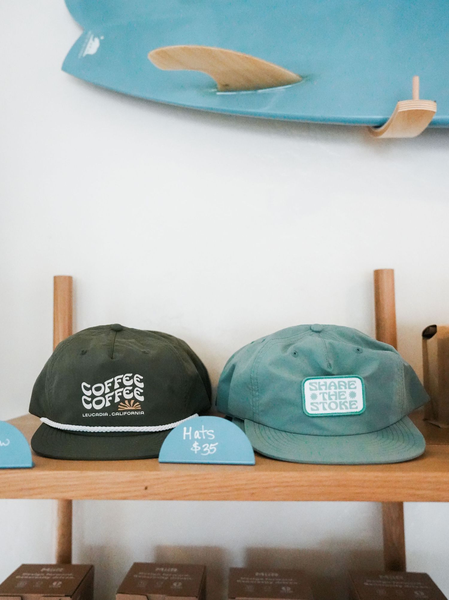 Three baseball caps displayed on a wooden shelf, featuring coffee-themed branding in green and blue colors.