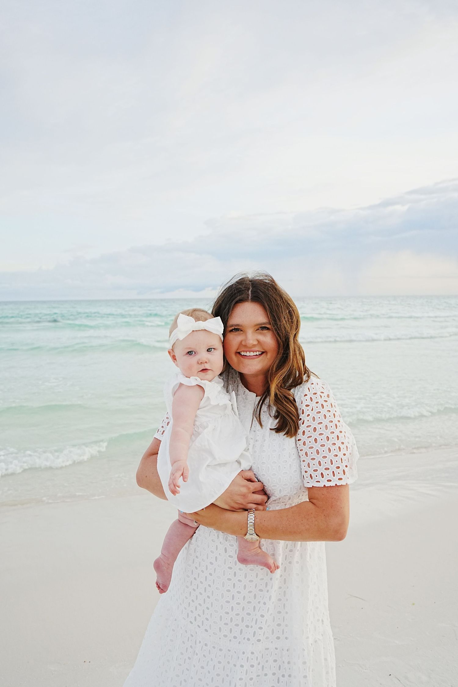 A parent and baby in white outfits share joyful moments together on a sunny beach with turquoise waters.