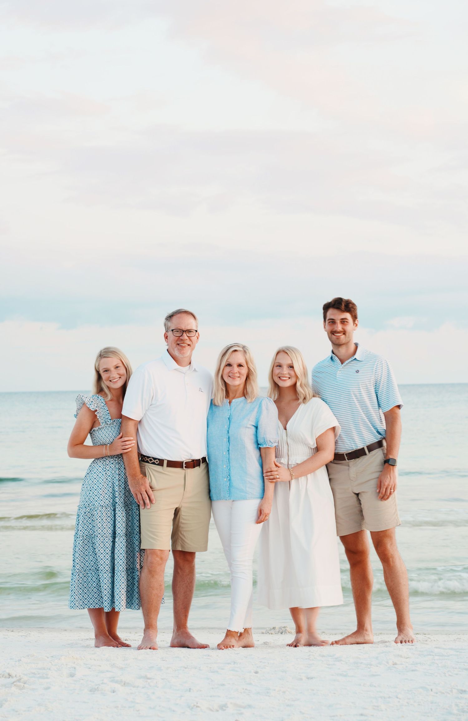 Family group in coordinated beach attire poses together on white sand beach at sunset with ocean in background.