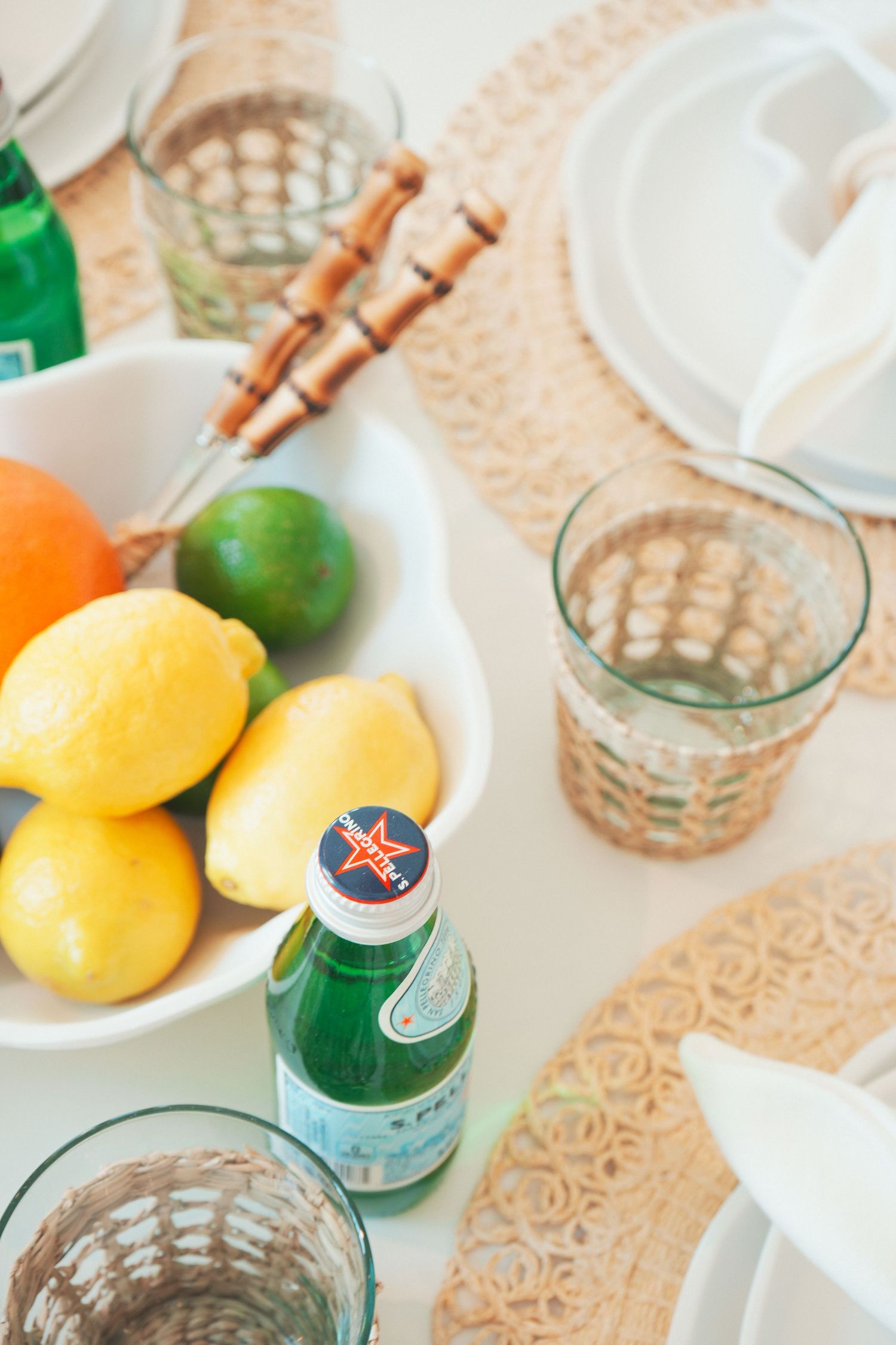 Fresh citrus fruits in a white bowl with sparkling water glasses and copper utensils on a beige patterned tablecloth.