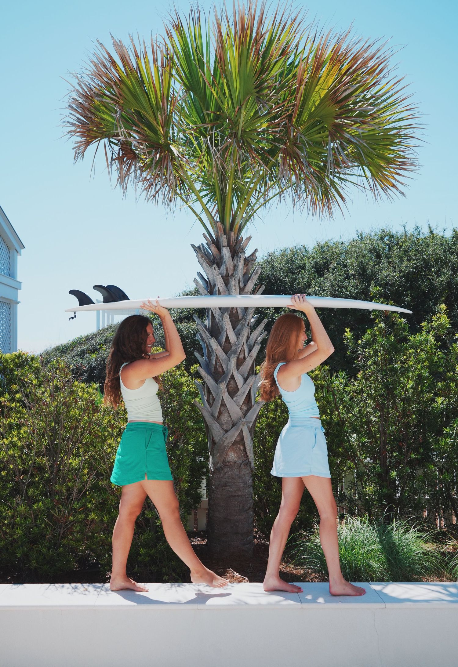 Two people in colorful summer outfits holding a surfboard overhead by a palm tree.