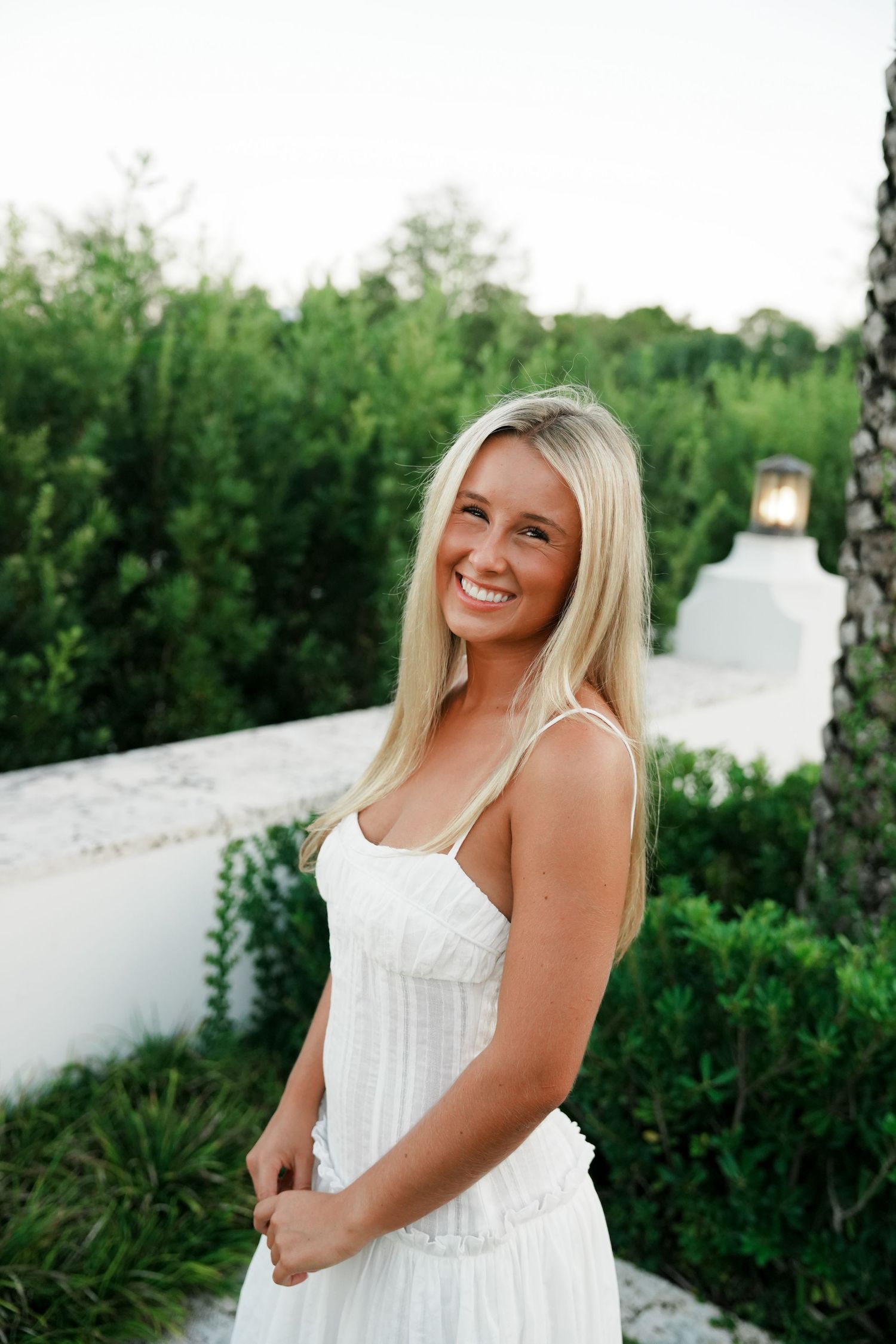 A person in a white sundress poses outdoors against a backdrop of lush greenery and white walls.