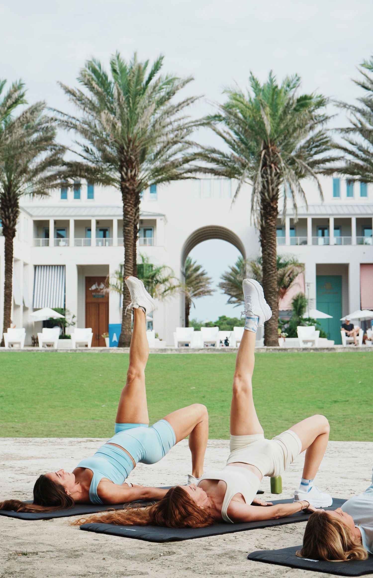 Two women practice relaxing yoga poses on yoga mats outdoors with palm trees and buildings in the background.