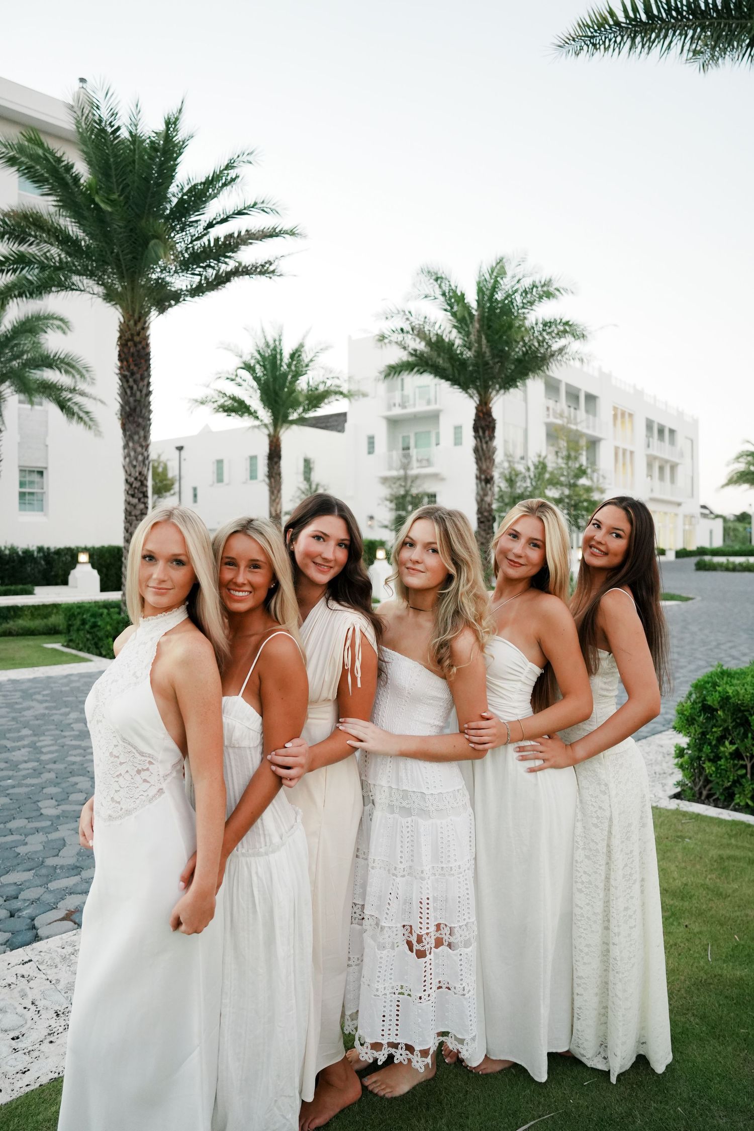 A group wearing coordinated white dresses poses together on a lawn with palm trees and white buildings in the background.