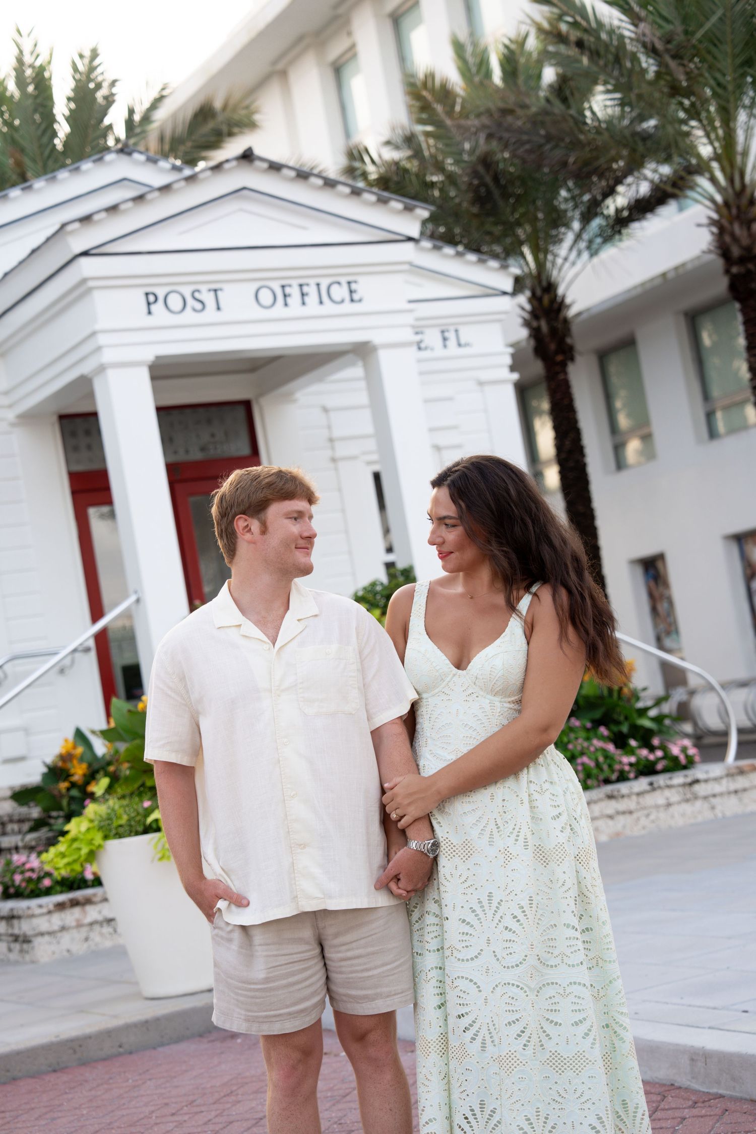 A smiling couple holds hands in front of a white Post Office building with columns and palm trees in Florida.