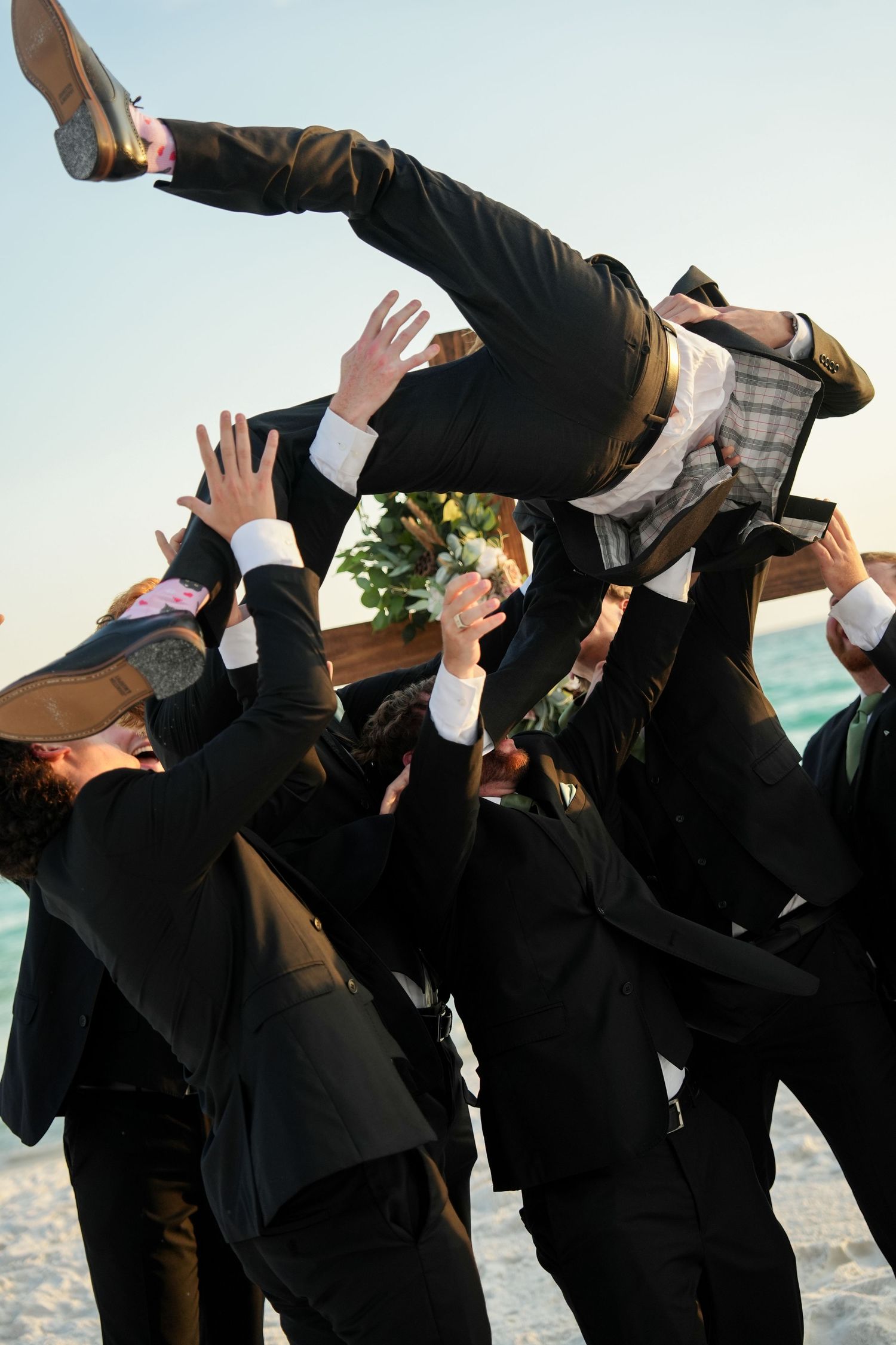 Groomsmen in black suits tossing the groom into the air on a beach during a wedding celebration.
