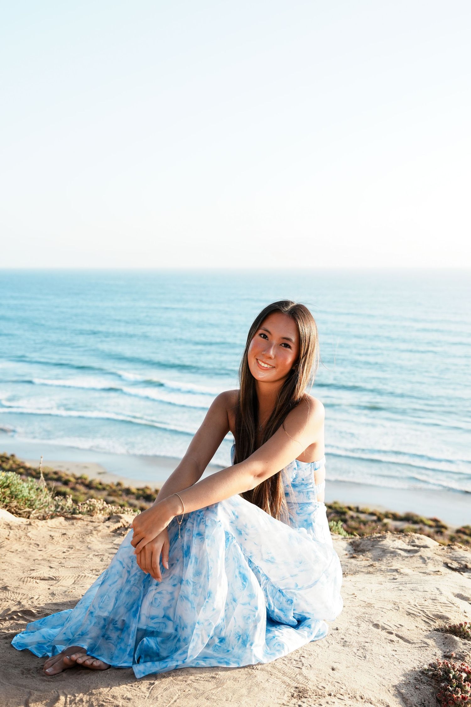 Young woman in blue floral dress sitting on sandy cliff overlooking ocean waves, smiling at camera.