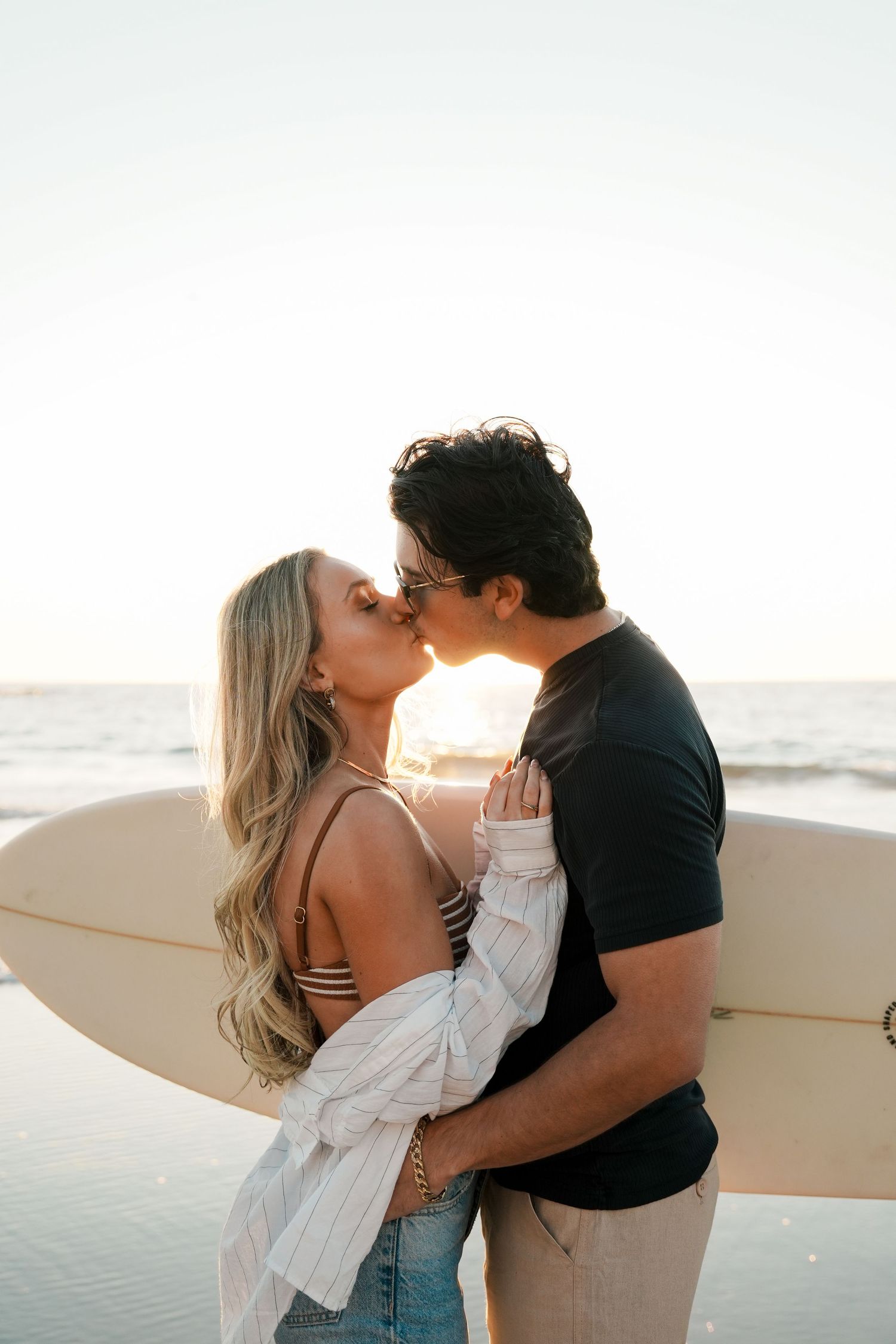 A romantic couple embraces on the beach at sunset with a surfboard nearby creating a silhouette against the ocean.