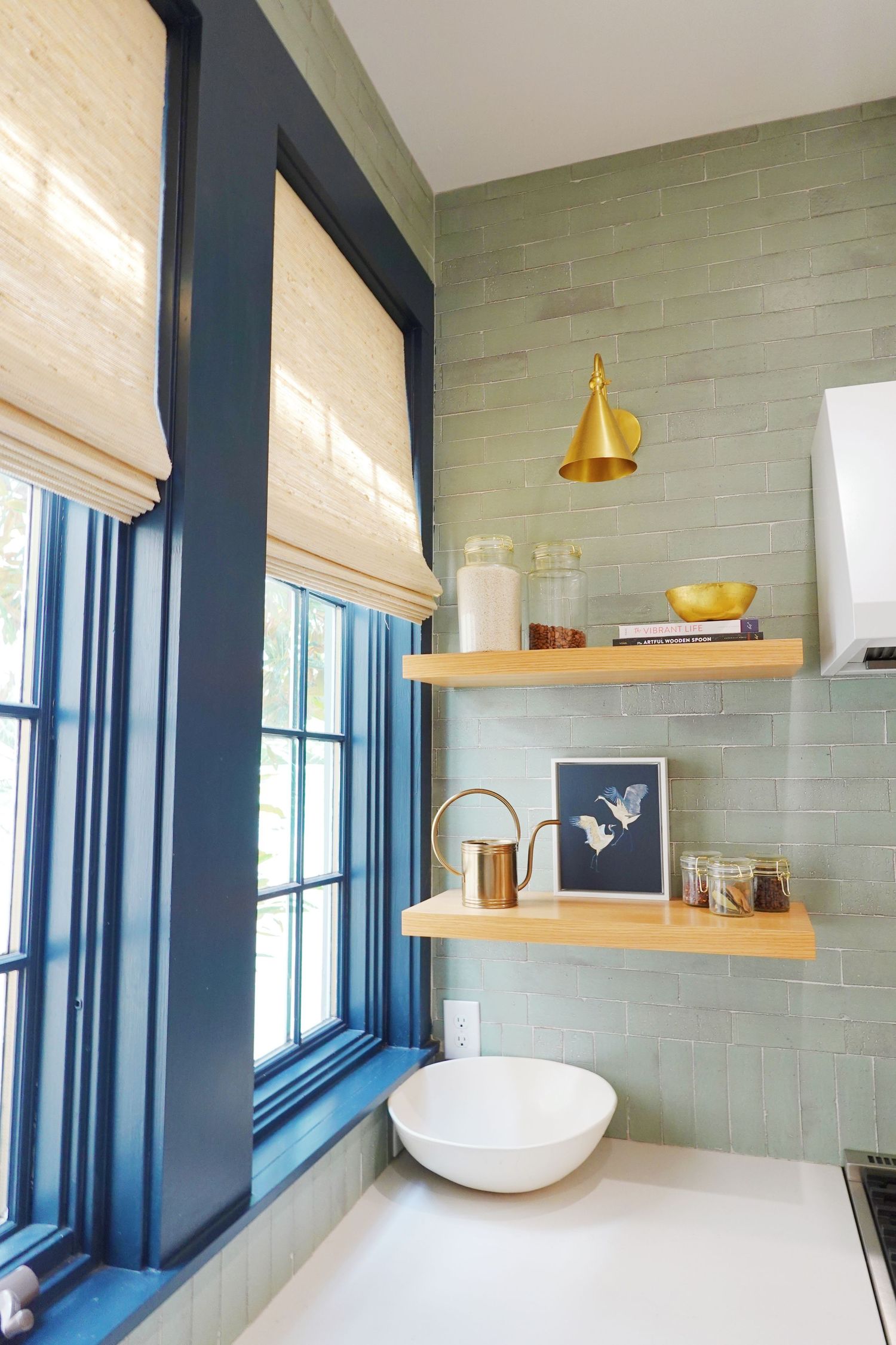 Coastal bathroom with navy trim, bamboo shades, and floating wooden shelves against sage green tile.