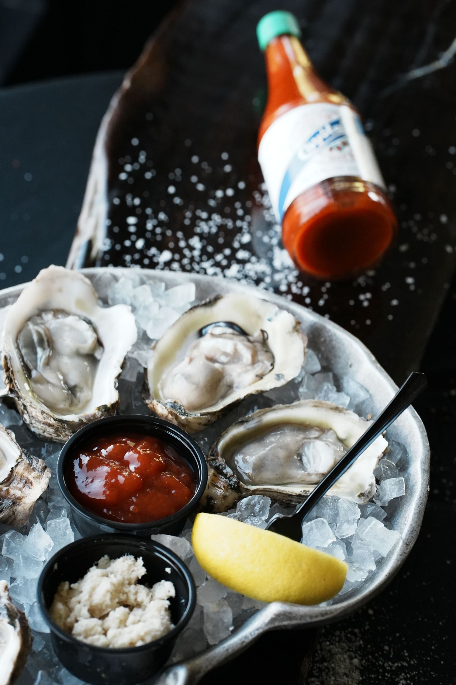 Fresh raw oysters on ice served with cocktail sauce, horseradish, and lemon wedges on a dark surface.