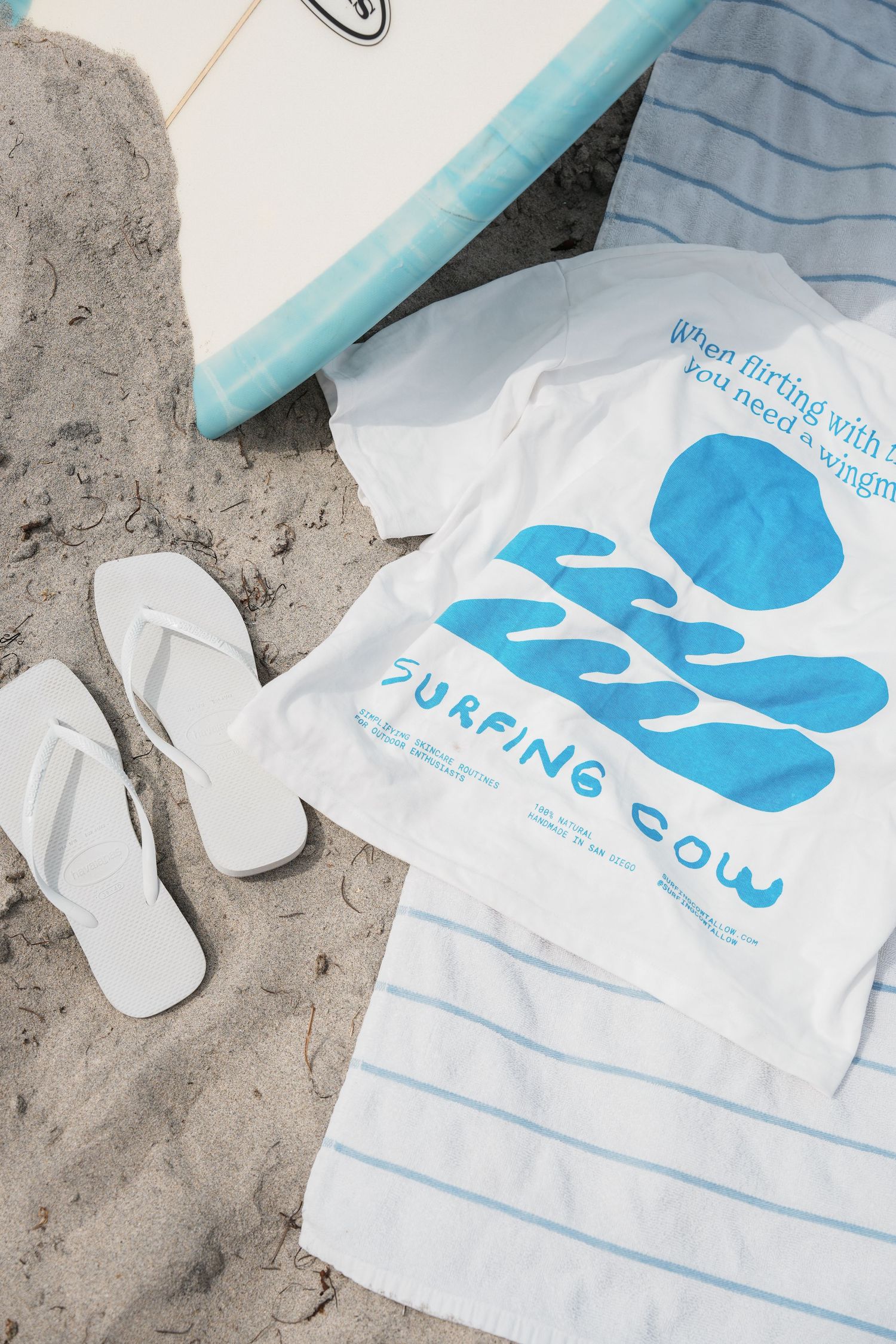 White t-shirt with blue surfing-themed design and graphics lying on sandy beach.