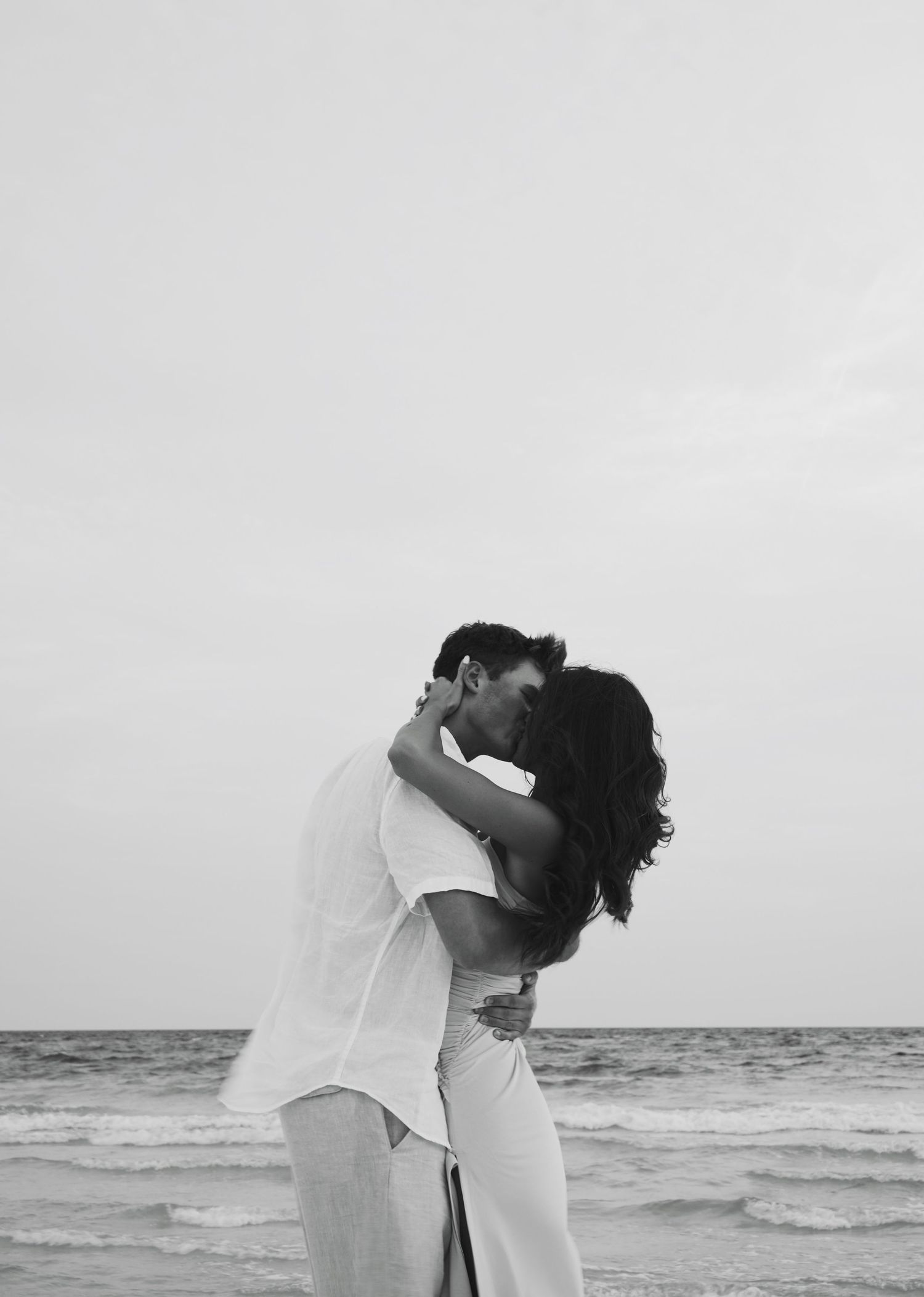 A couple in white clothing shares an intimate moment together on a beach at sunset, creating a romantic silhouette.
