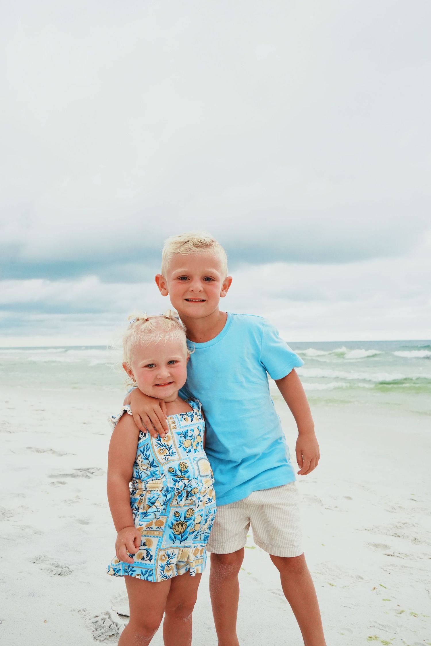 Two young children in summer outfits smile while posing together on a cloudy beach with ocean waves in the background.