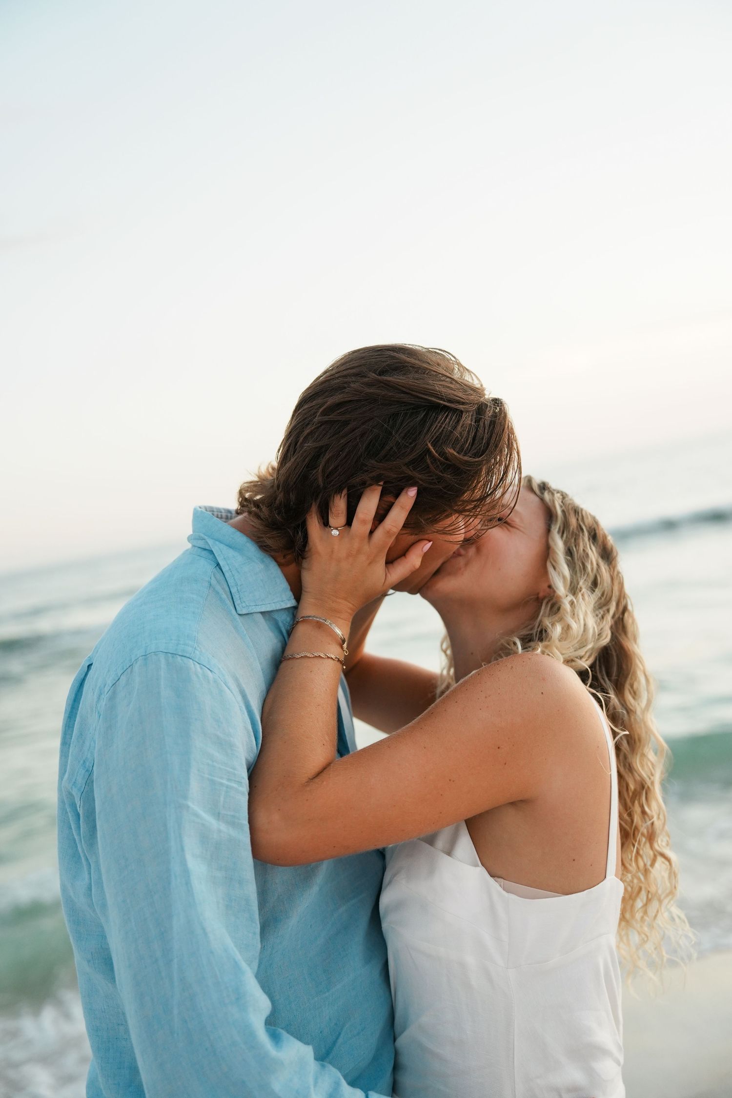 A romantic couple shares a tender kiss on the beach at sunset with ocean waves in the background.
