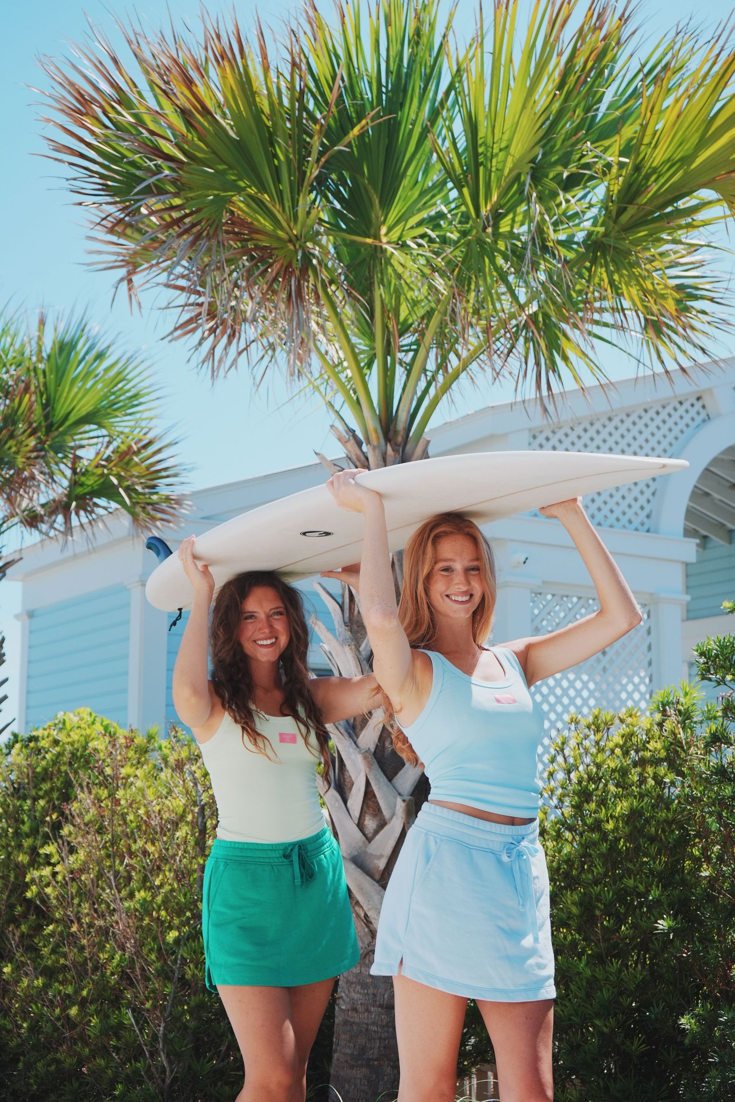 Two friends holding a surfboard overhead near tropical palm trees on sunny day.