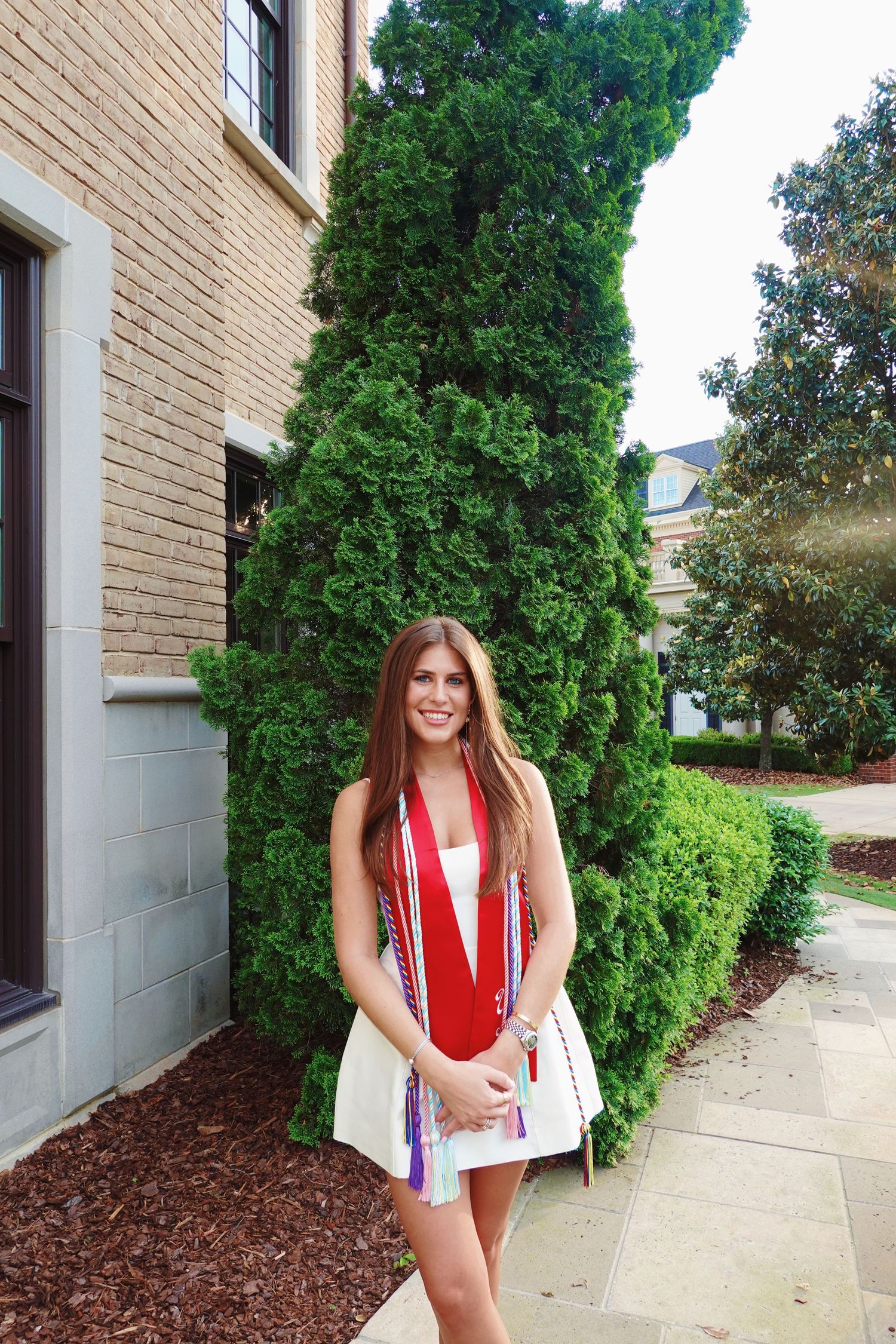A person in a red and white swimsuit poses against a beige brick wall with green shrubs.