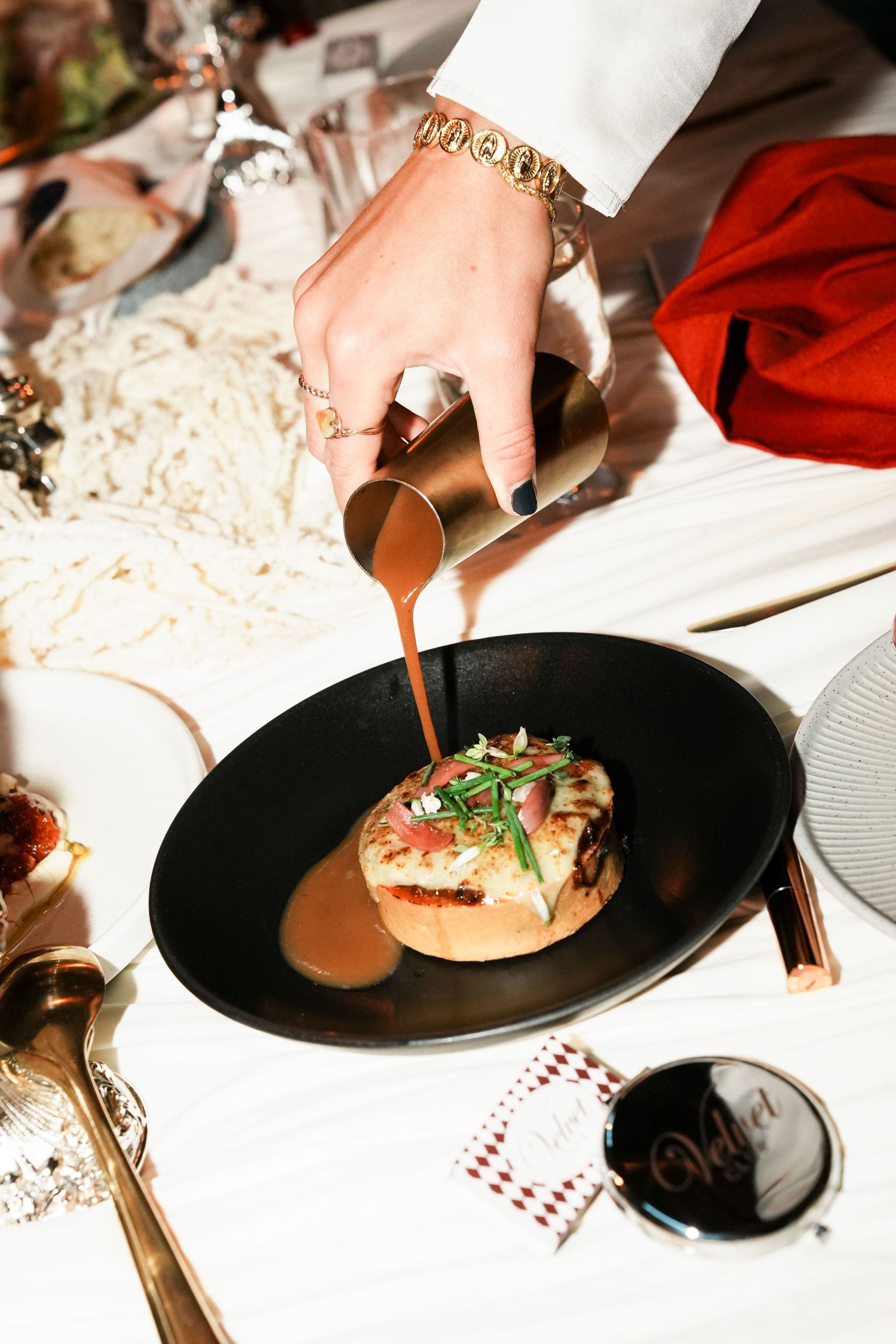 A hand pouring rich brown sauce from a copper pitcher onto an elegant gourmet dish garnished with greens in a black plate.