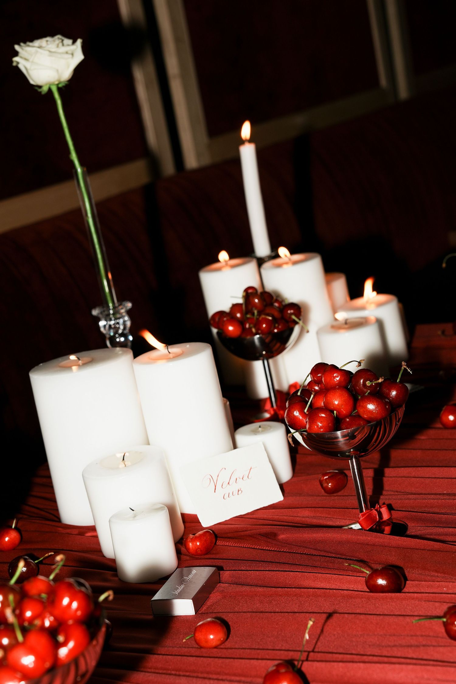 Elegant wedding centerpiece with white pillar candles, fresh cherries in silver bowls, and a single white rose on a red table.