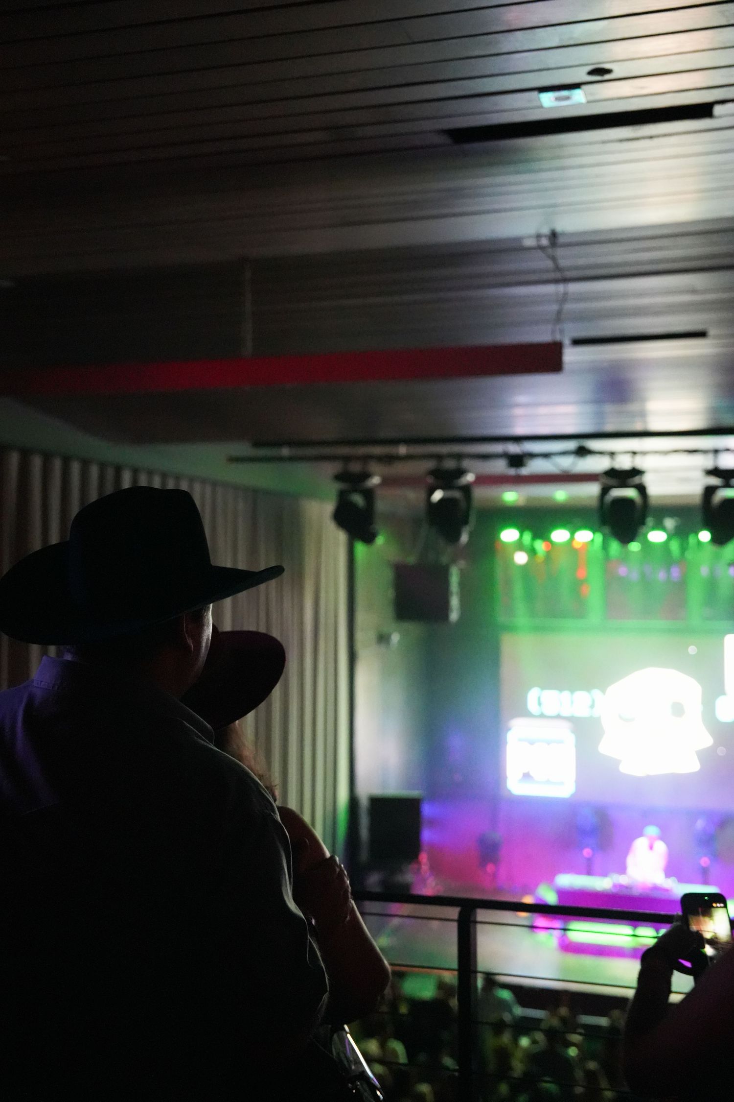 Silhouetted figure performs karaoke in a dimly lit bar with colorful stage lighting and neon signs in the background.