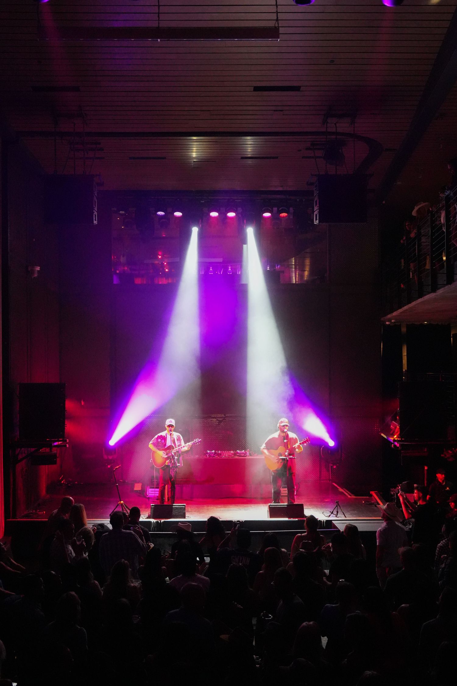 Concert stage illuminated with dramatic purple and white spotlights shining down on performers as crowd watches in silhouette.
