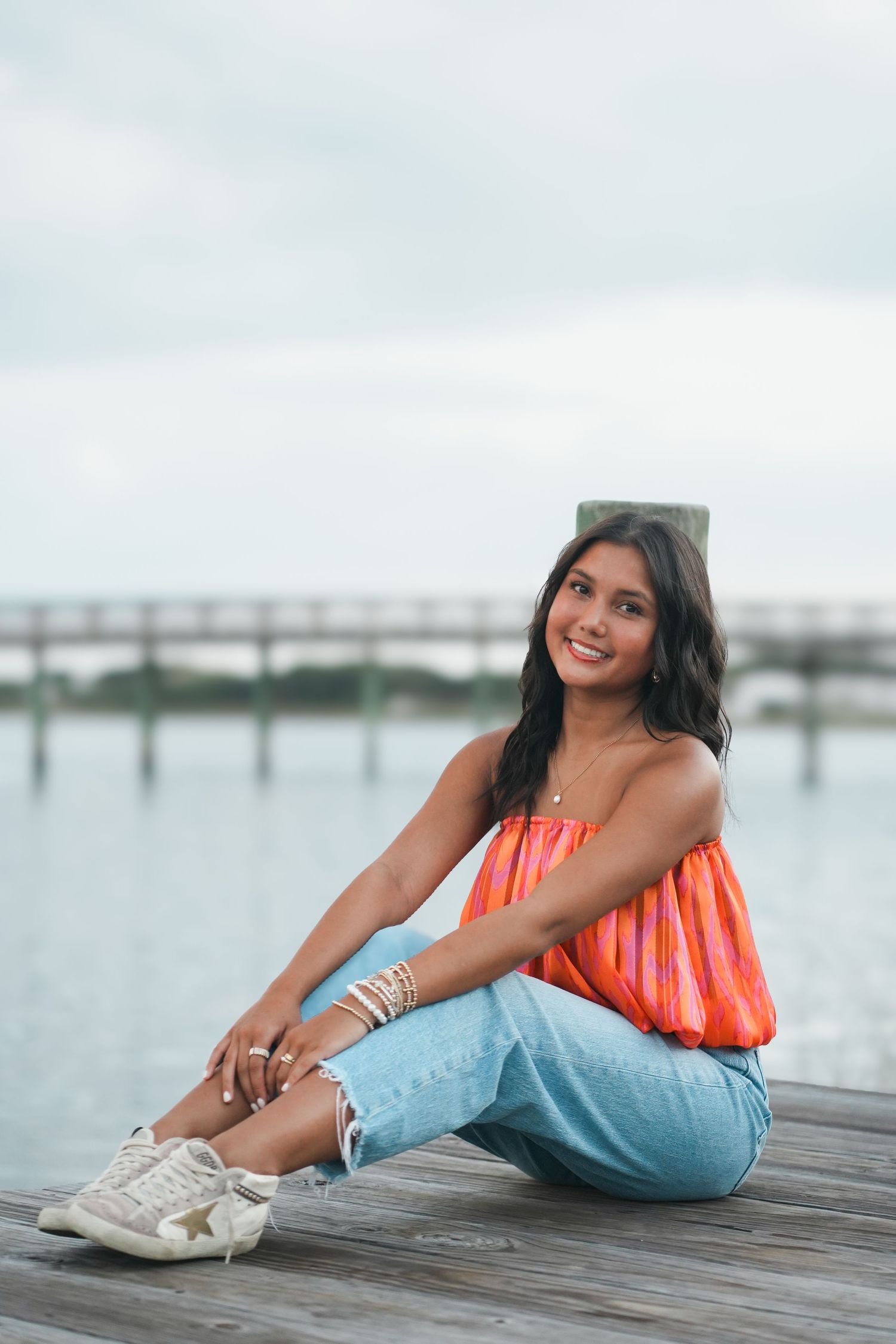 A casual summer outfit featuring an orange top and jeans while sitting on a wooden dock by the water.