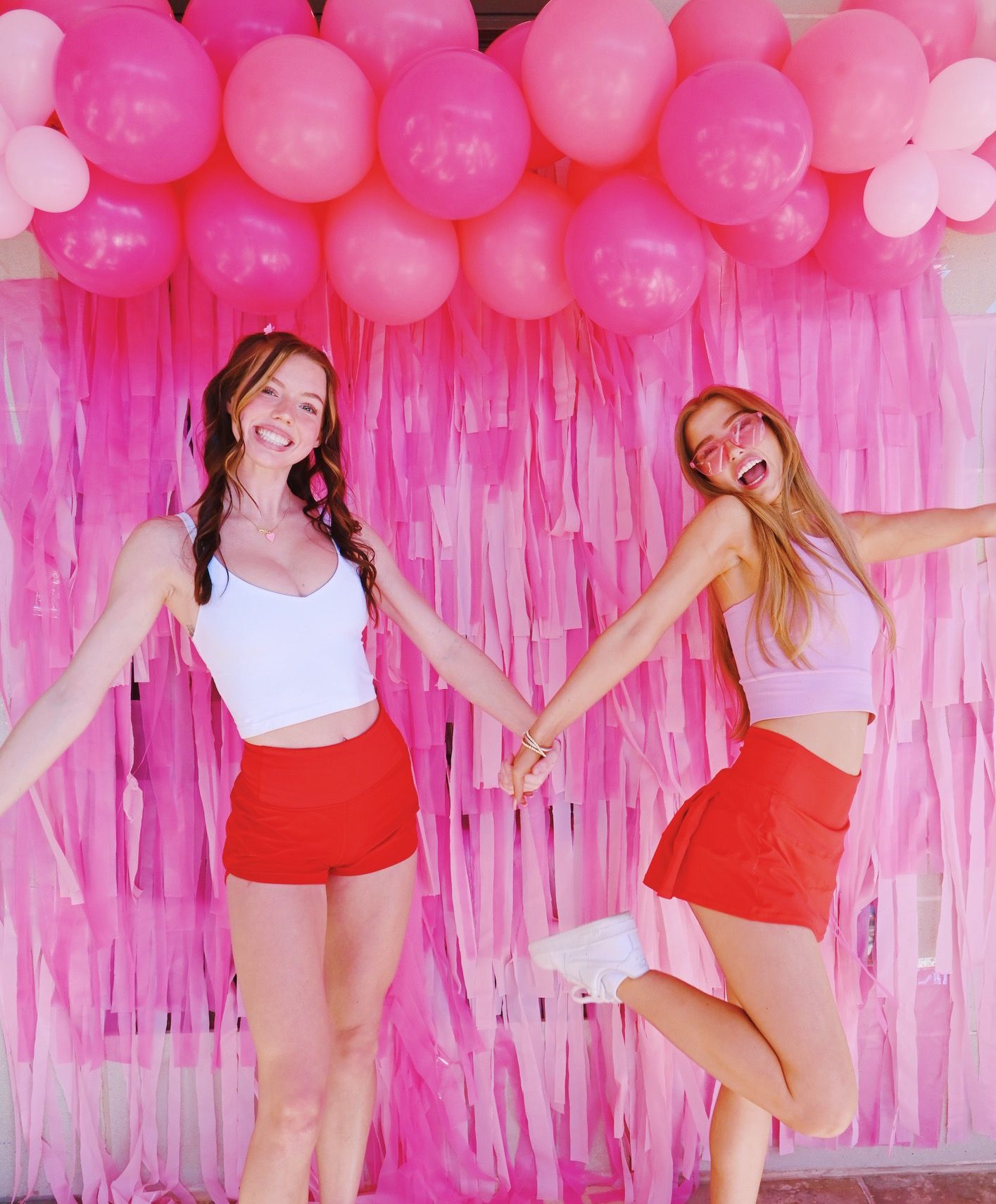 Two friends posing playfully against a pink balloon and streamer backdrop at a party.