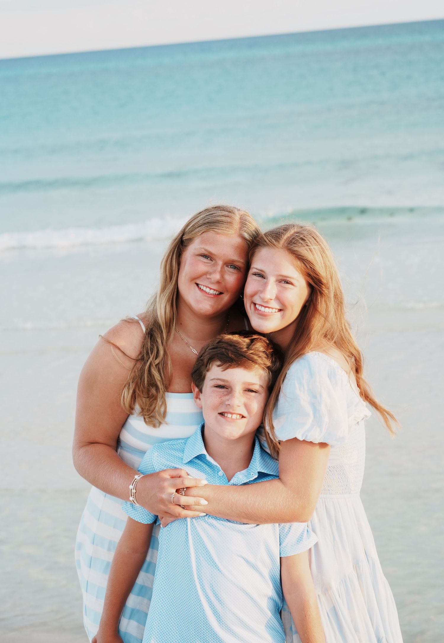 A family in white and light blue outfits poses together on a sunny beach with turquoise water in the background.