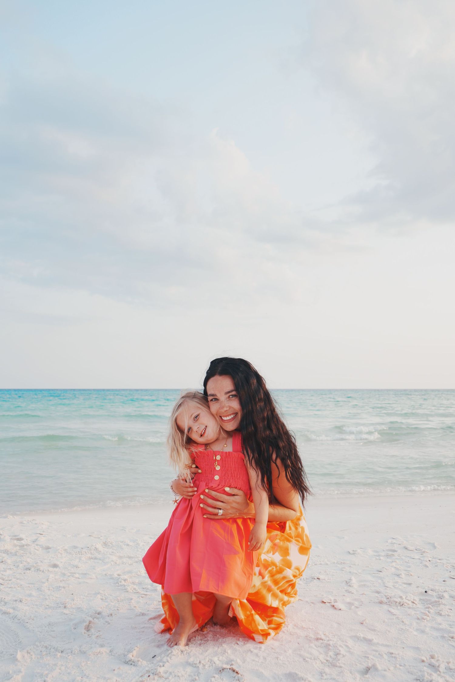 Two people embrace on a white sandy beach with turquoise waters and cloudy sky in the background.