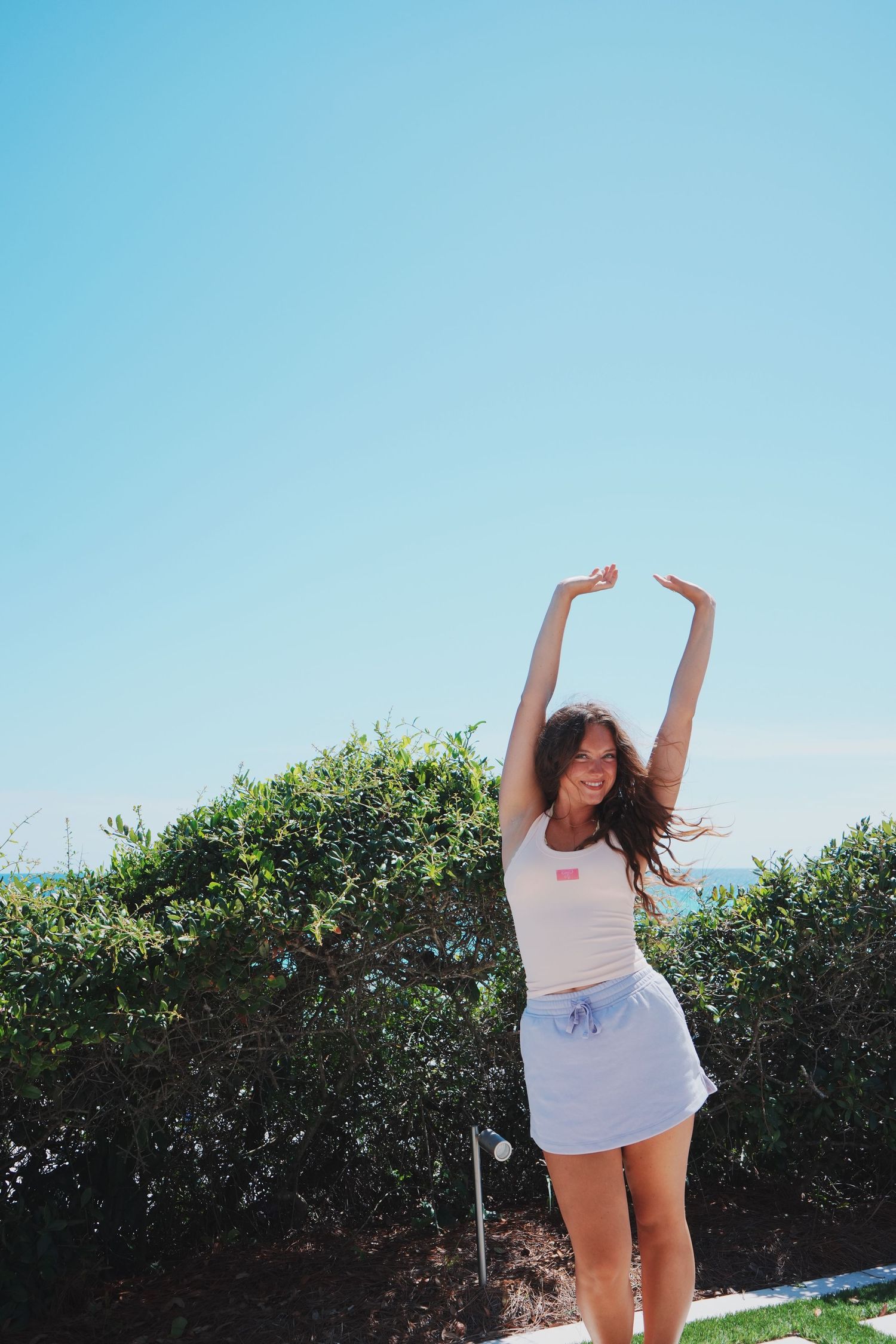 A person in summer attire stretching joyfully with arms raised against a bright blue sky and ocean view.