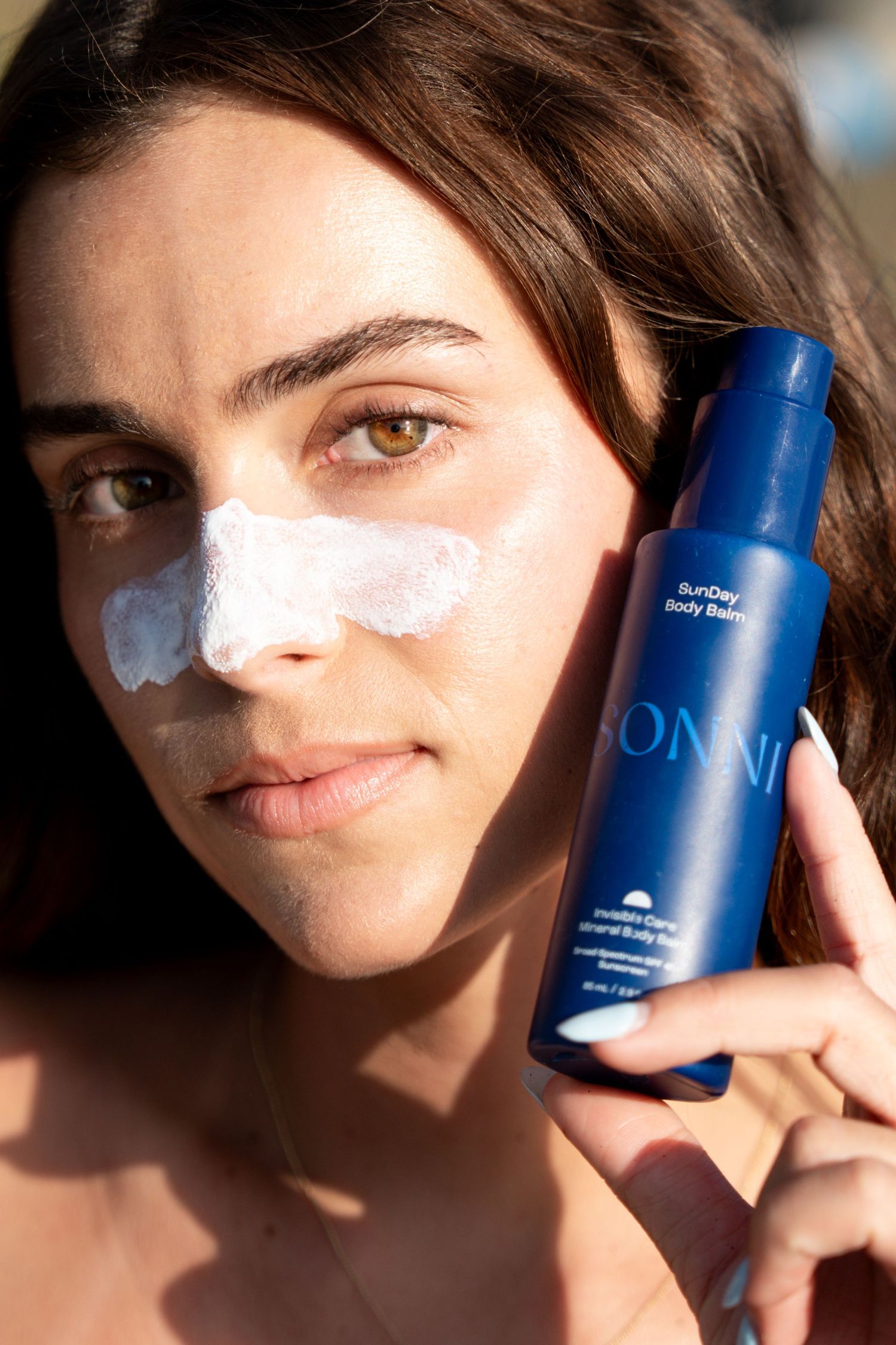 Woman applying SONNI SunDay Body Balm sunscreen to her face, holding blue bottle outdoors in natural sunlight.