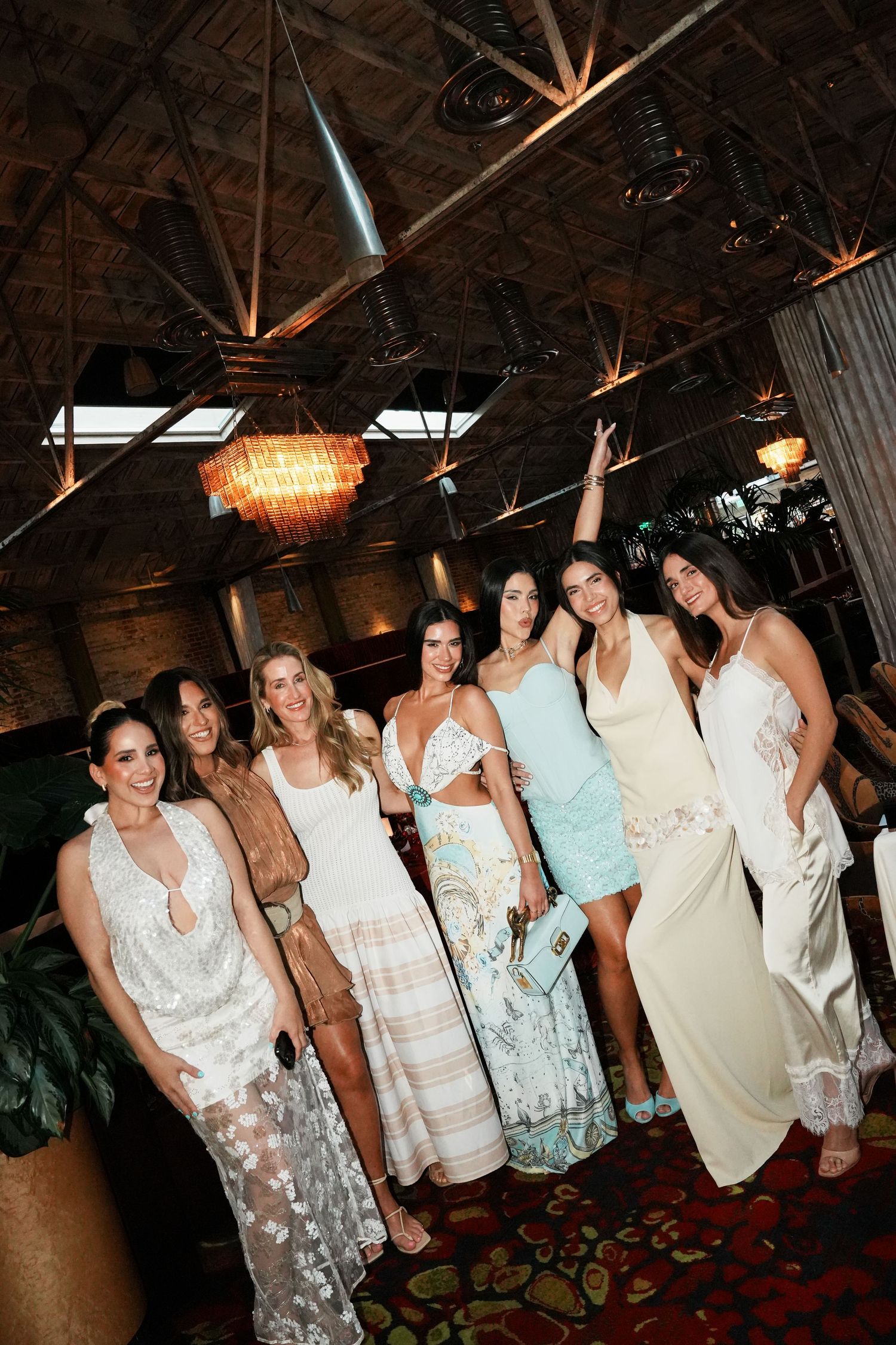 Group of seven women in elegant white and pastel dresses posing together at an upscale indoor event venue.