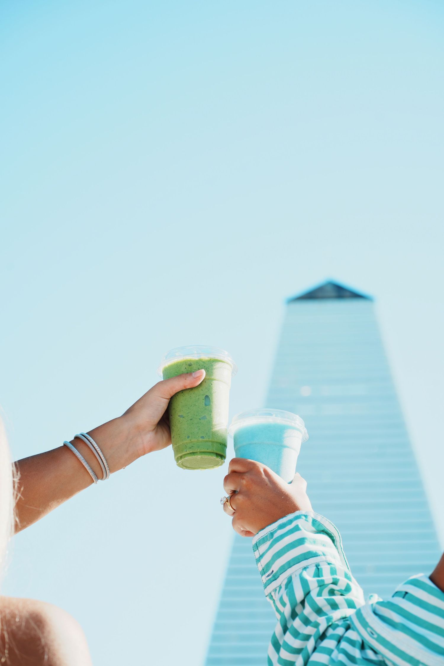 A person in a striped shirt holds up a green smoothie against a bright blue sky with urban buildings in the background.