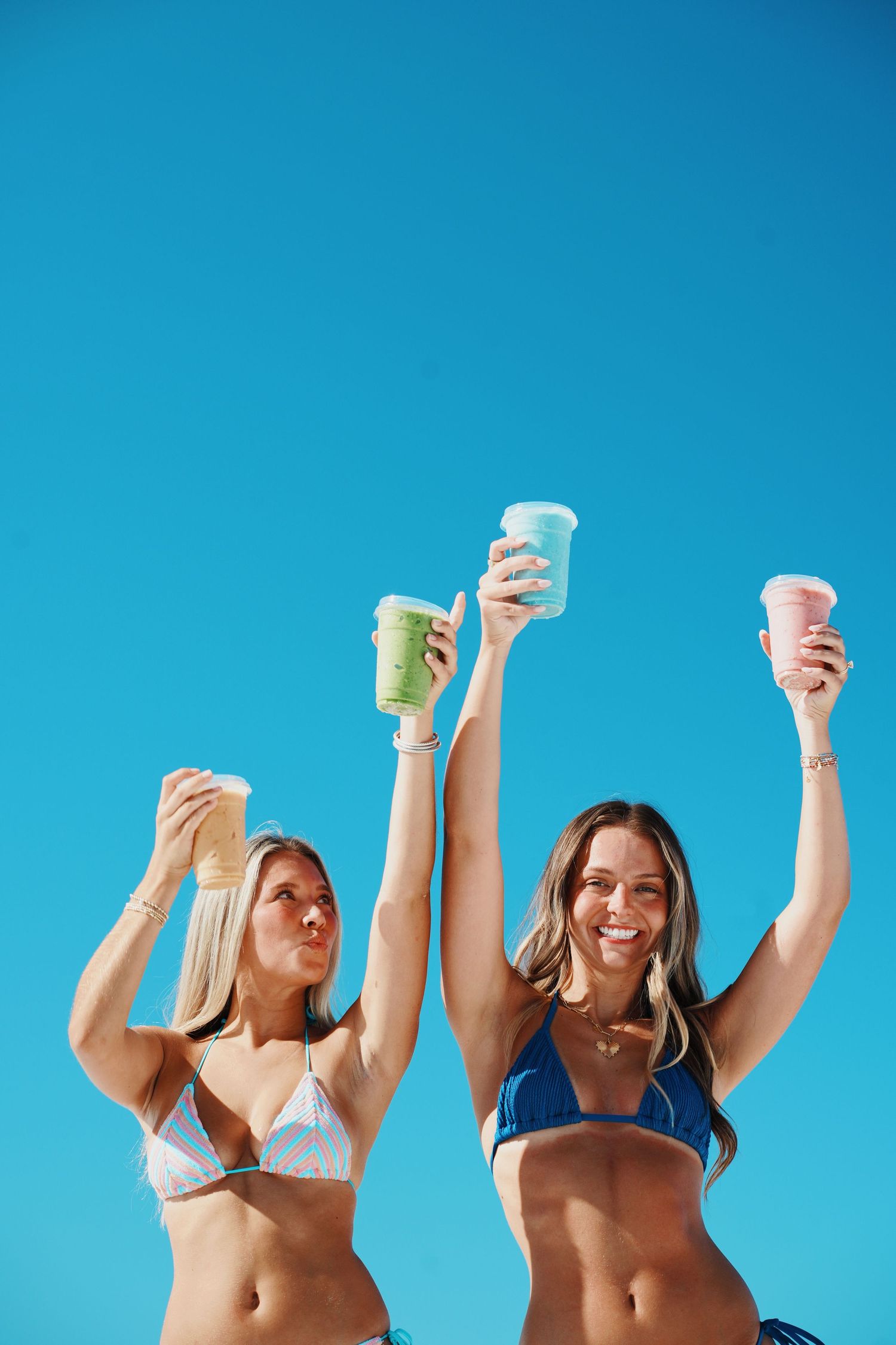 Friends raising colorful smoothies against bright blue sky in celebration.