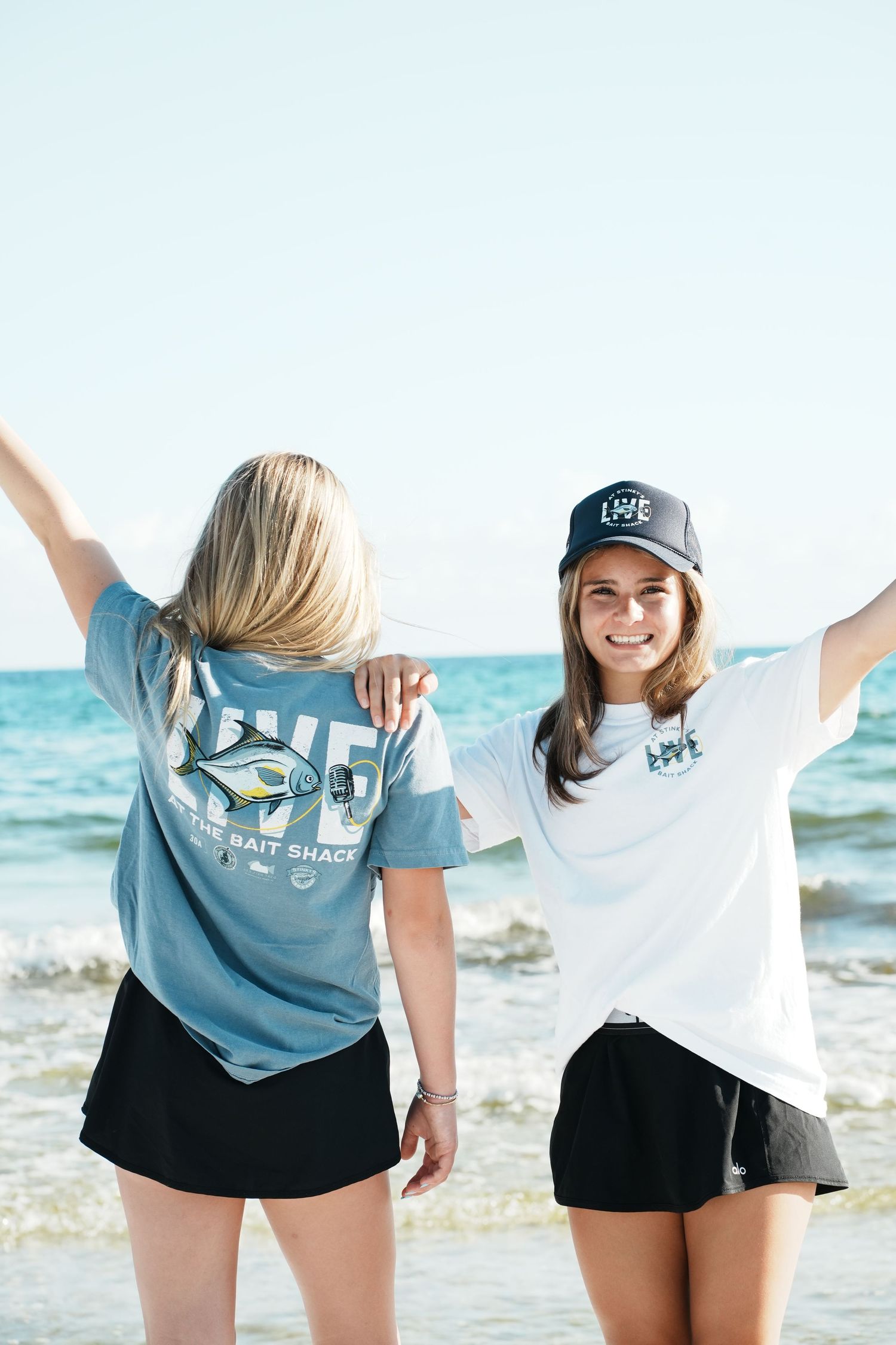 Two friends wearing casual beachwear and baseball caps pose with arms raised by the ocean on a sunny day.