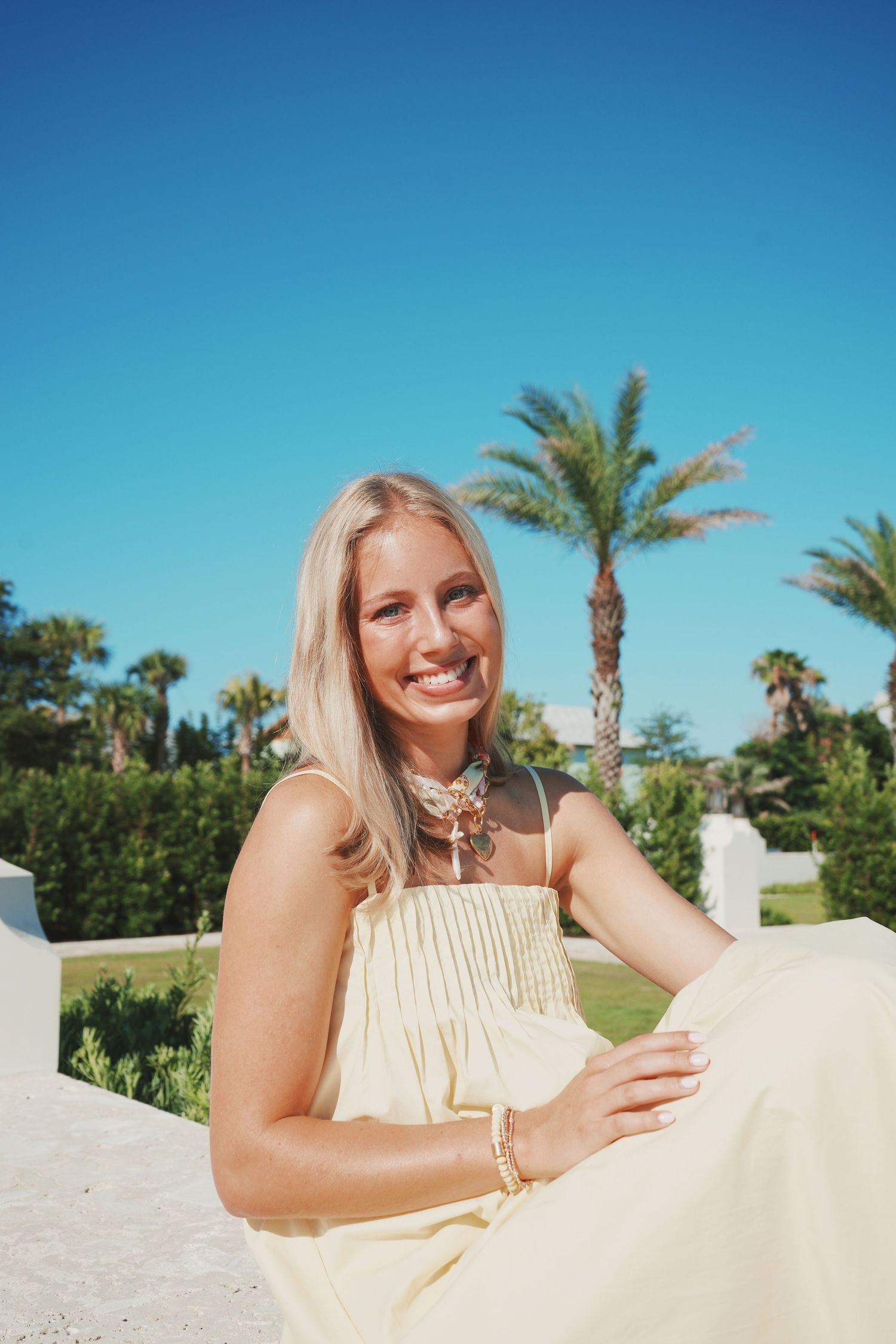 Woman in a cream sundress relaxes at a tropical resort with palm trees and blue sky in the background.