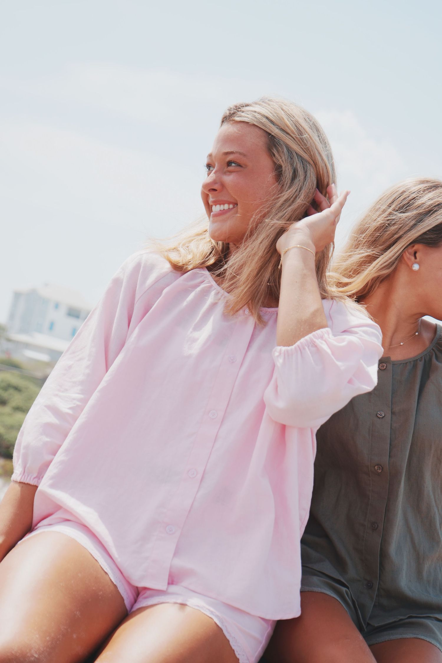 Two people in casual summer clothing sit together outdoors, with one wearing a pink blouse and smiling towards the horizon.
