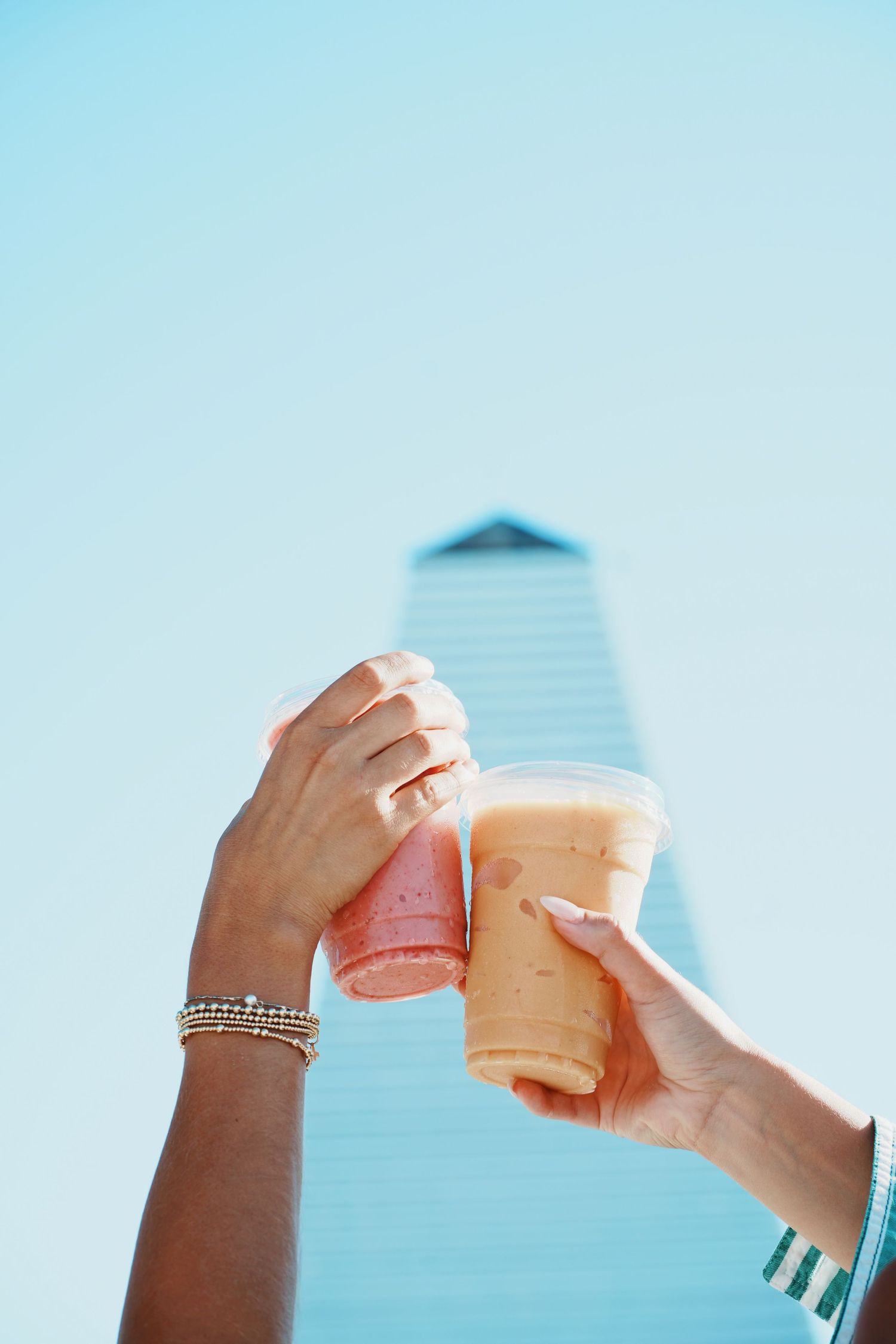 Two hands cheering with colorful iced drinks against a blue sky with a building in the background.